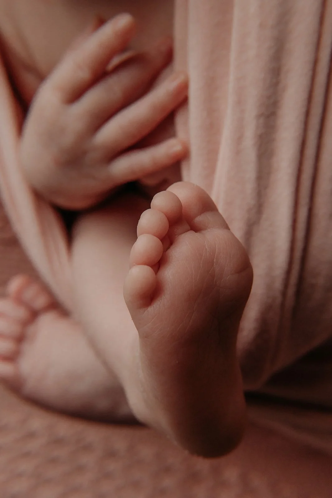 Close-up of a newborn baby's tiny foot being held gently, with part of the baby's hand visible resting on their chest, all wrapped in soft pink fabric.