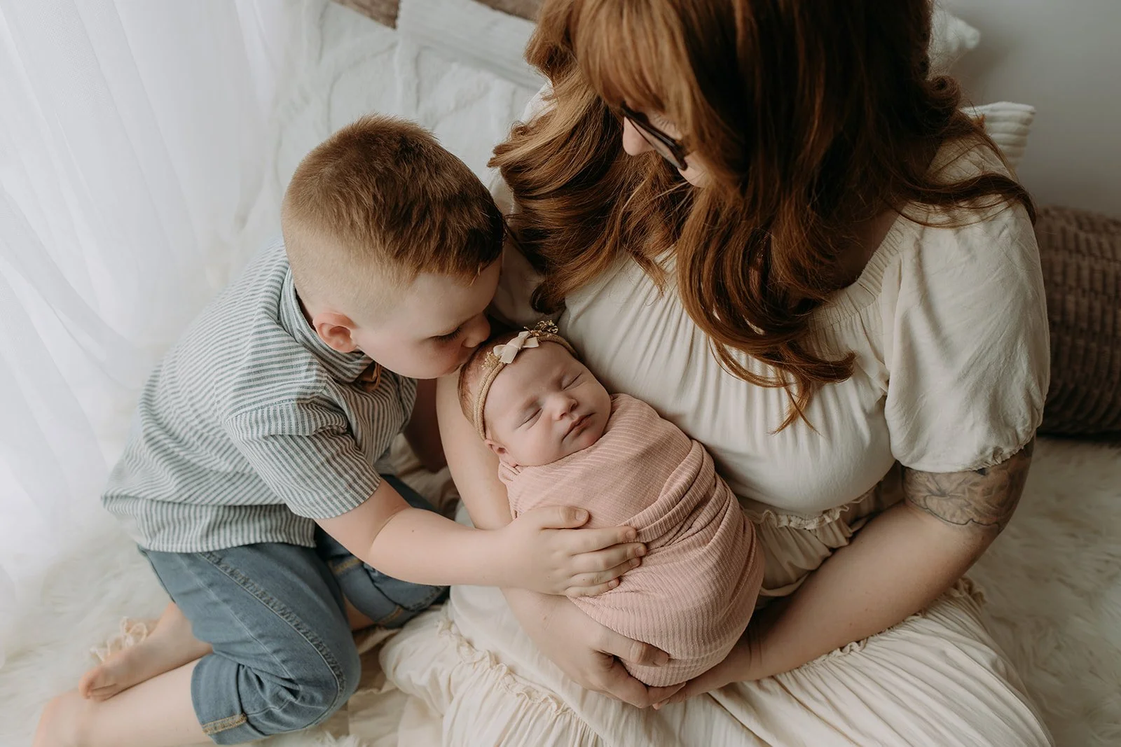 Young boy kissing a newborn baby wrapped in pink on a woman’s lap