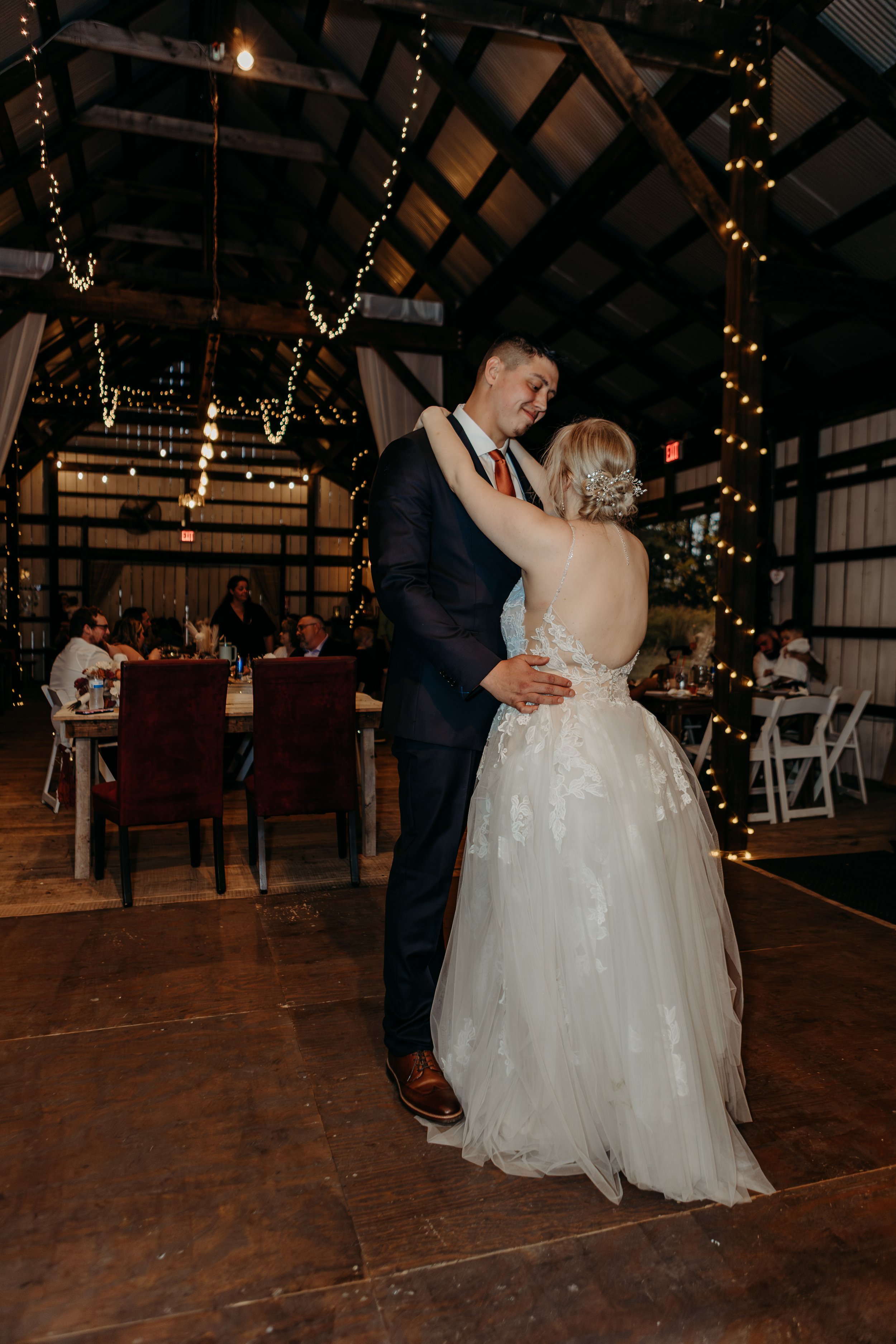 Bride and groom dancing during wedding reception in barn with fairy lights, guests seated in background.