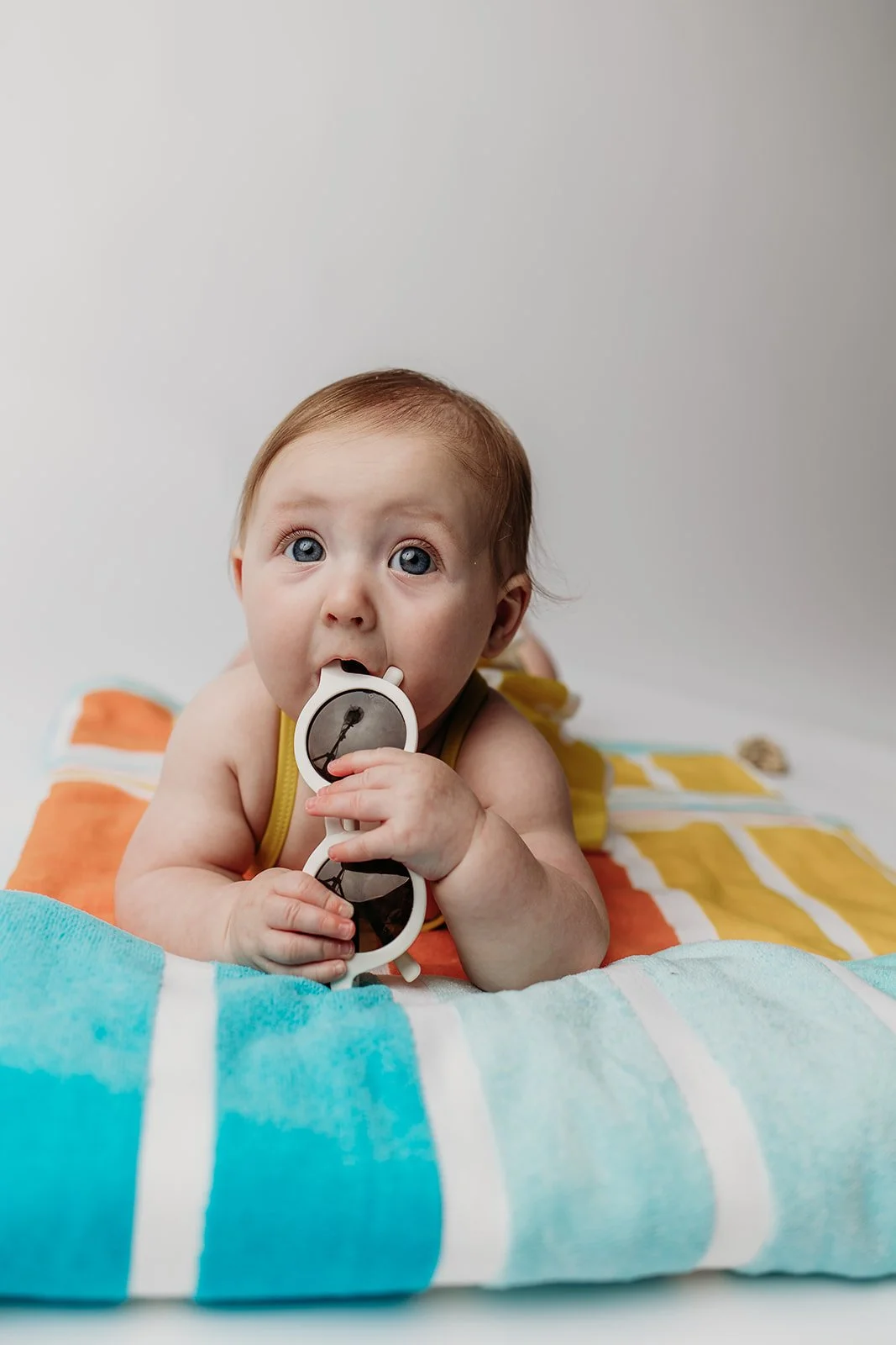A baby lying on a colorful striped towel, holding sunglasses, and looking at the camera