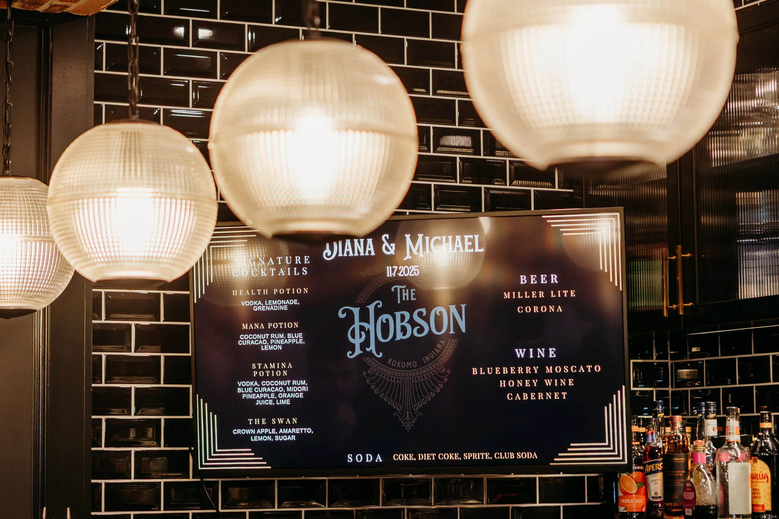 A bar menu screen displaying drinks and their ingredients, with hanging light fixtures above, against a black tiled wall.