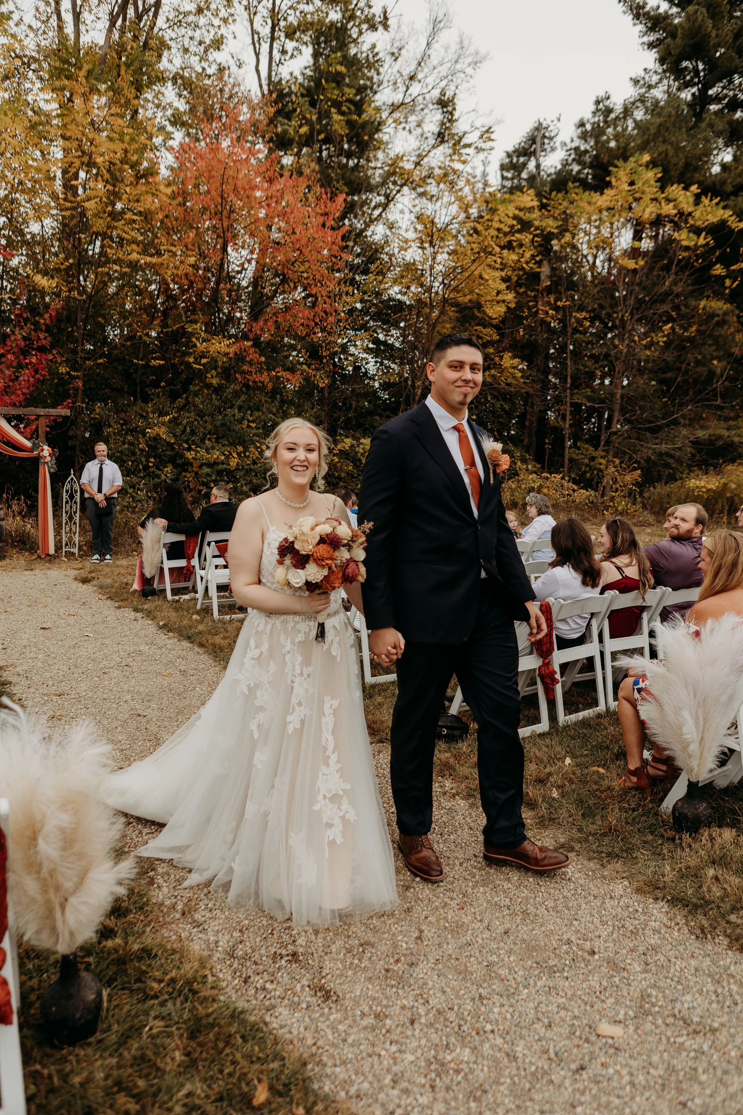 A wedding ceremony outdoors with a bride and groom walking down the aisle, surrounded by guests seated on white chairs, autumn trees with colorful leaves in the background, and decorated with white and beige accents.