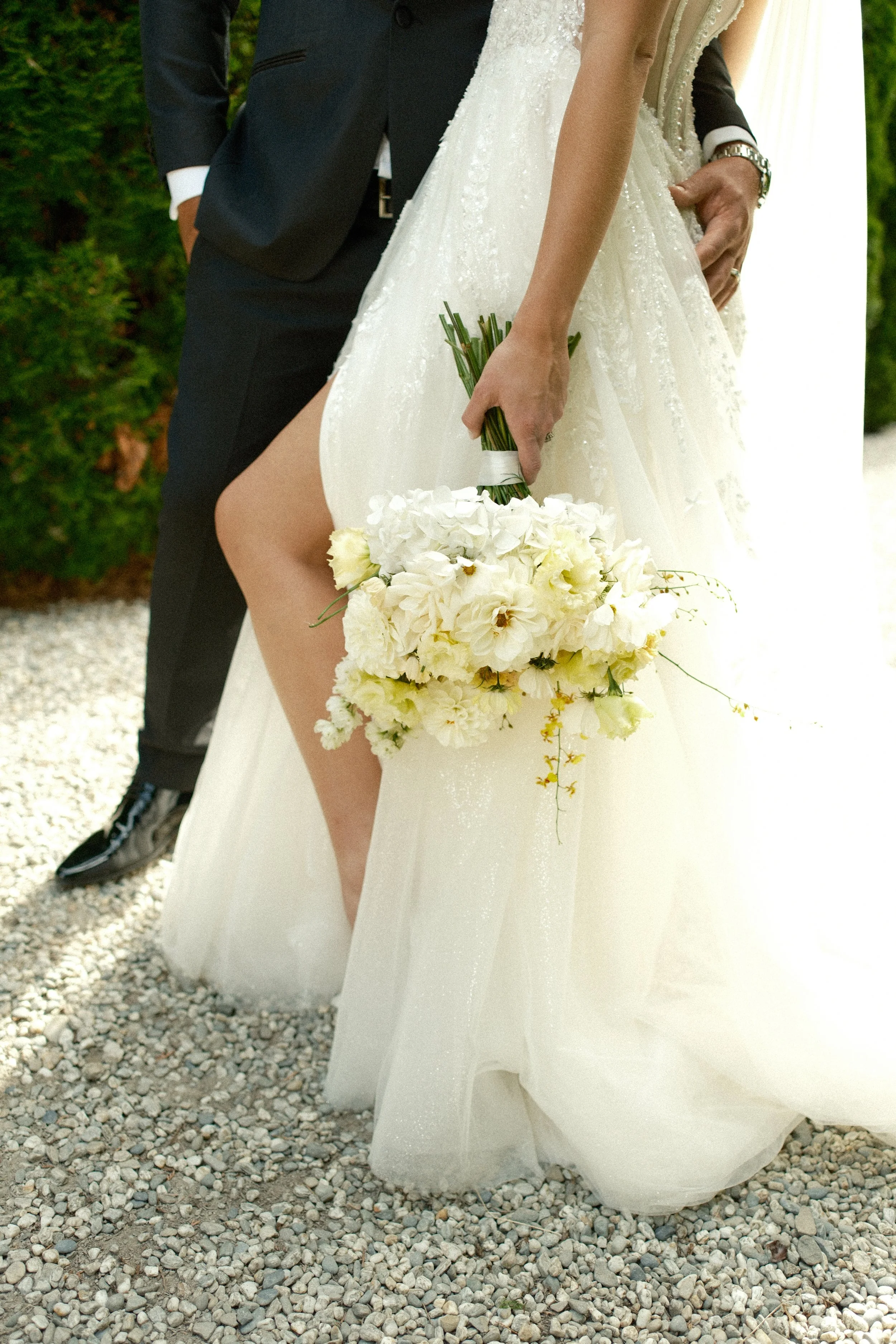 Bride in a wedding gown holding a bouquet of white flowers, standing next to groom in a tuxedo, on a gravel path outdoors.
