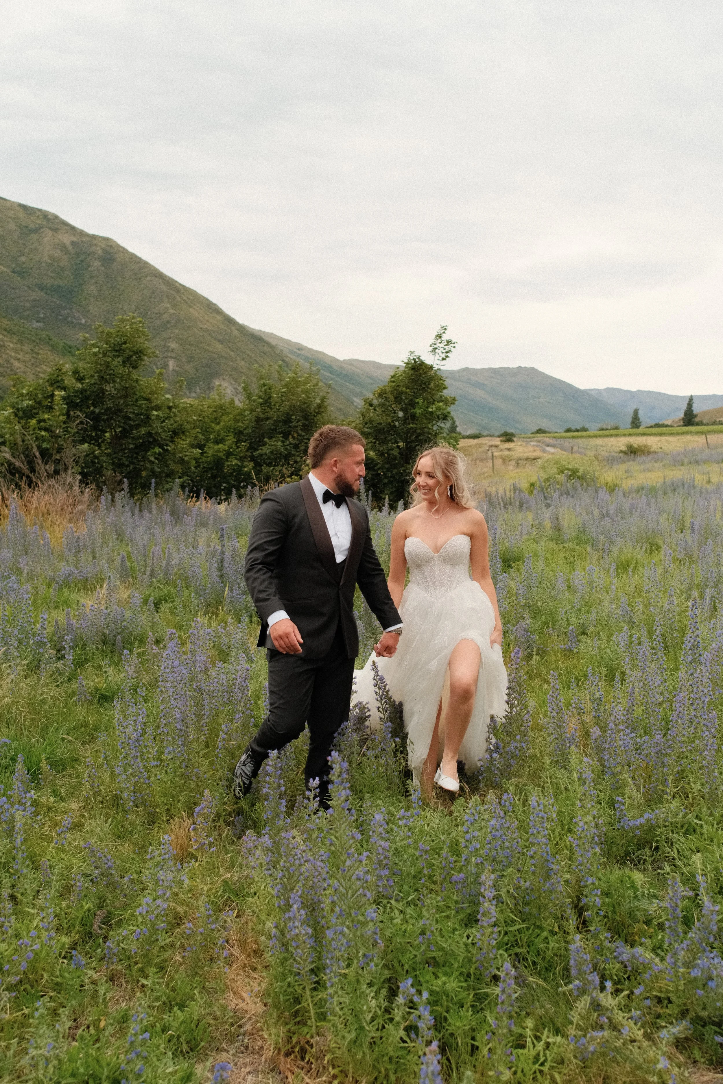 Bride and groom walking hand in hand through a field of purple flowers with mountains in the background.