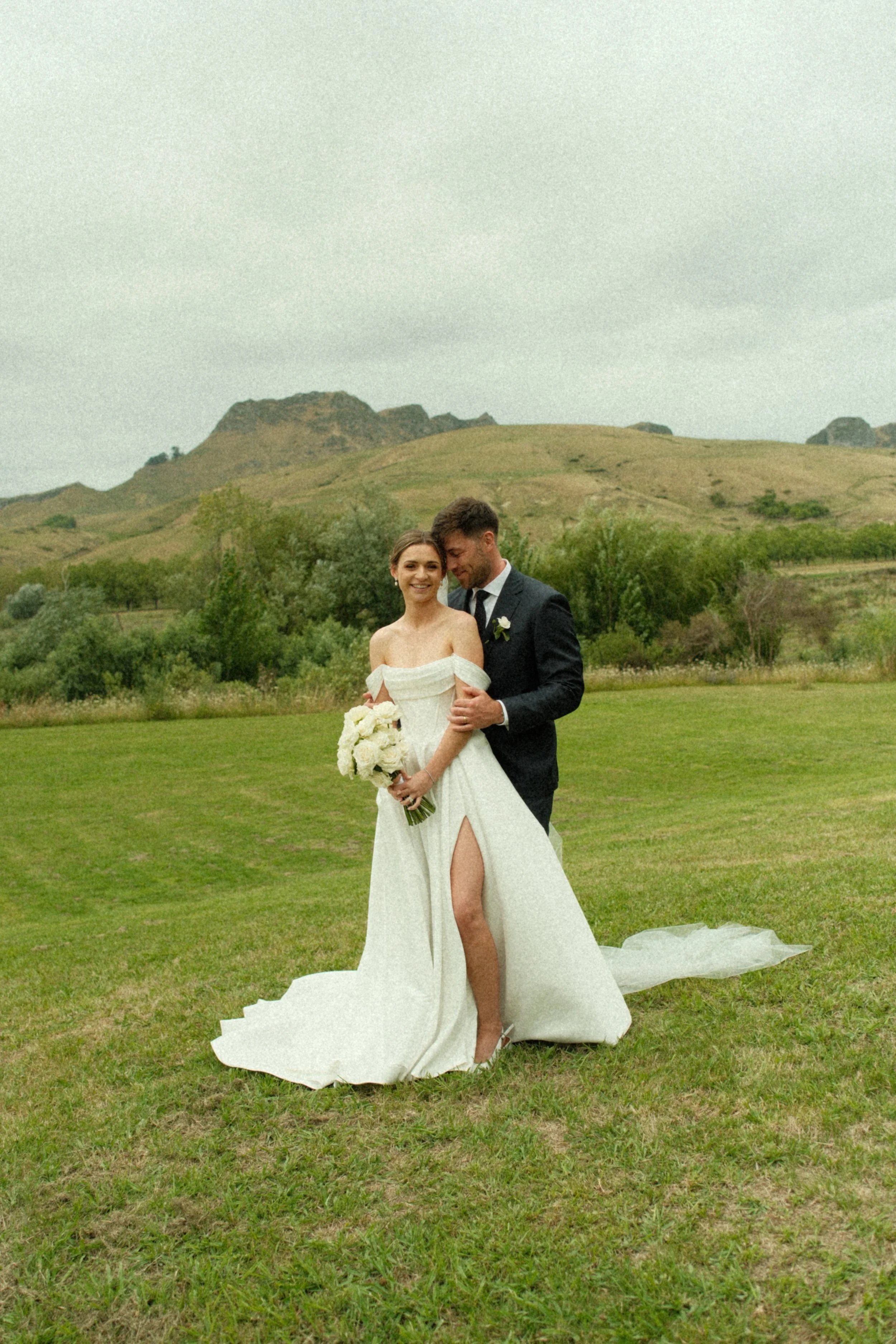 A bride and groom standing on a grassy field, smiling and embracing, with a mountain and cloudy sky in the background. The bride wears a white off-the-shoulder wedding dress with a high slit, holding a bouquet of white flowers. The groom wears a dark