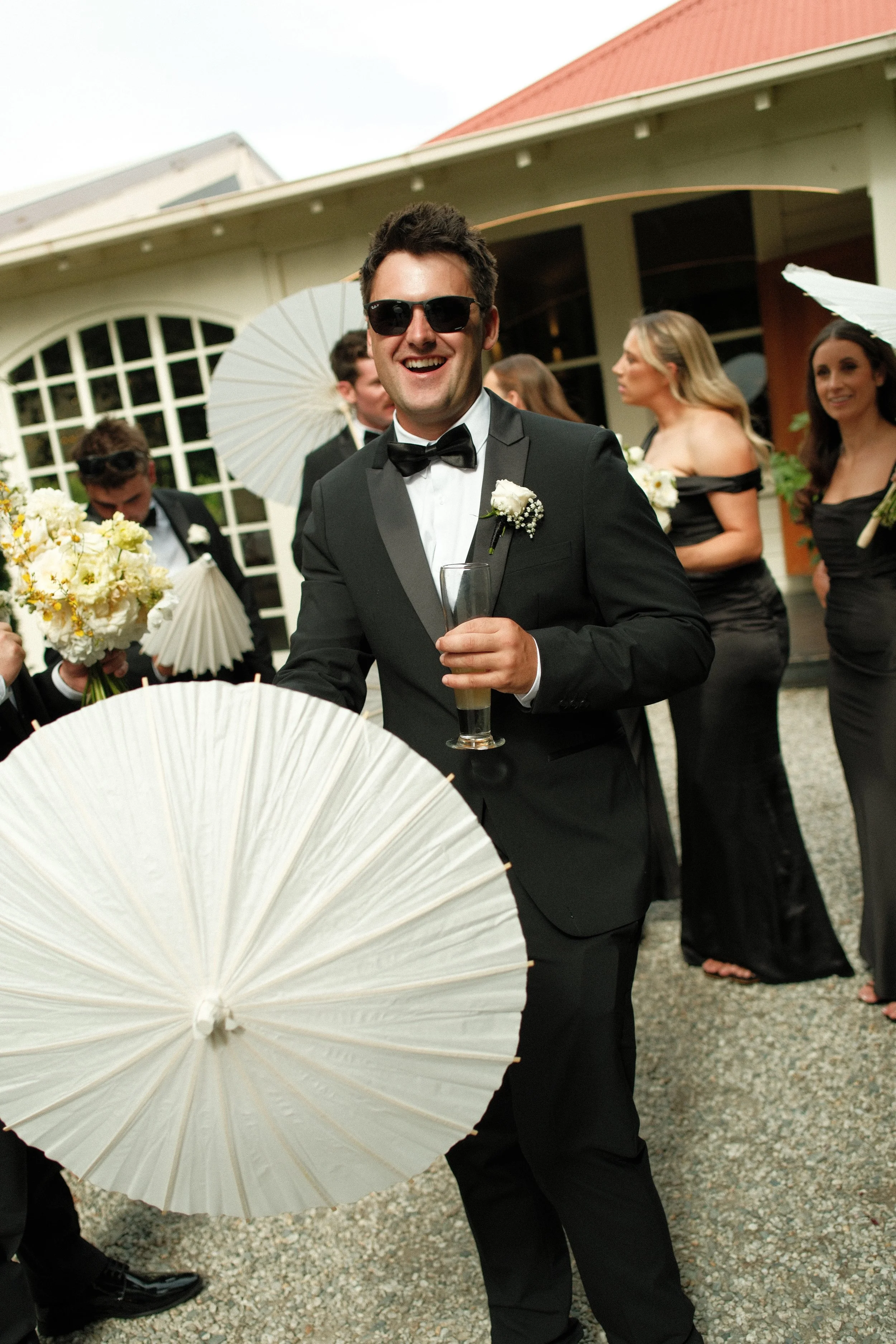 Man in tuxedo and sunglasses holding champagne glass at wedding reception with bridesmaids and groomsmen in black dresses and suits, holding bouquets and umbrellas, outside building.
