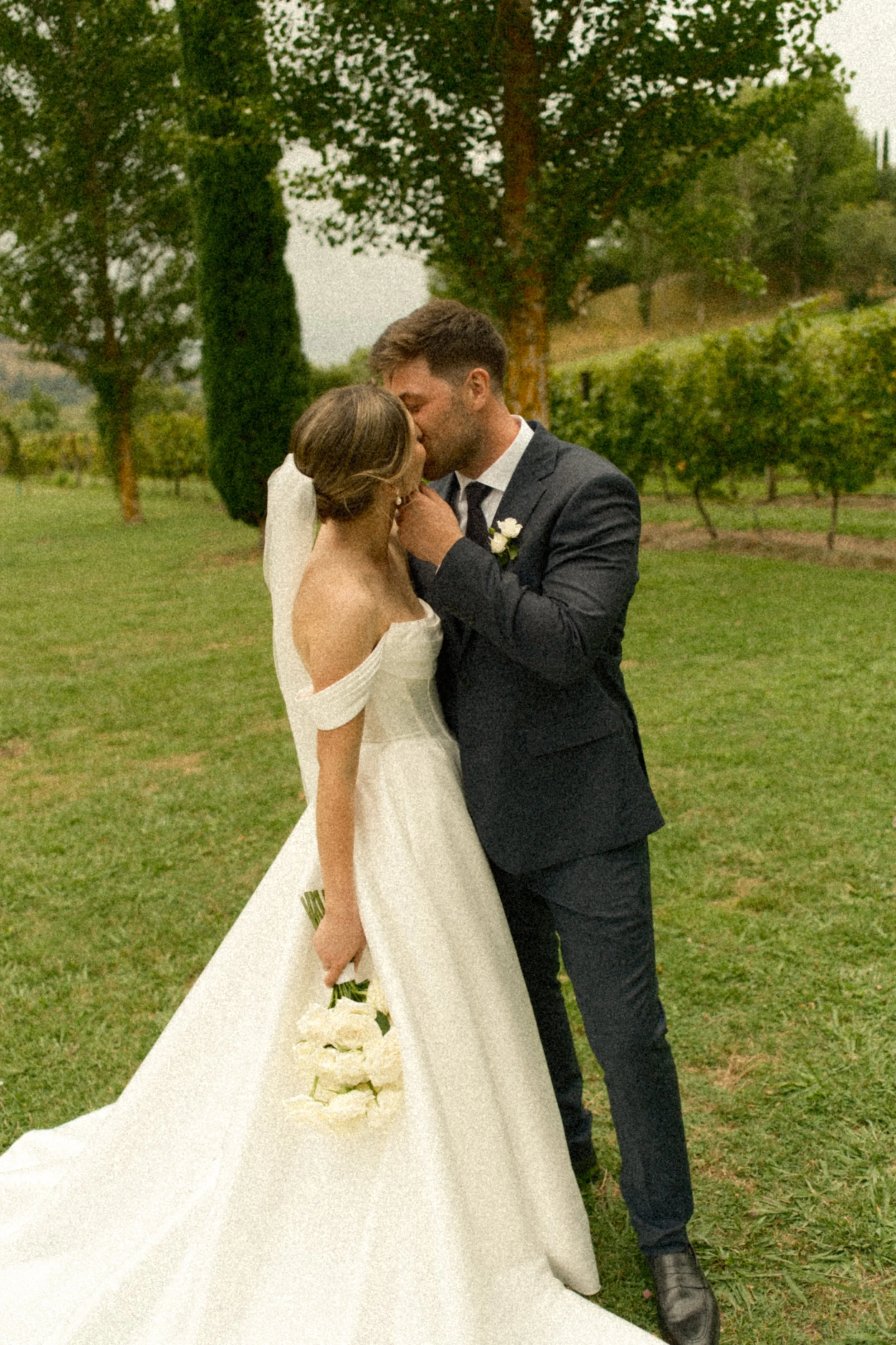 A bride and groom share a kiss outdoors on their wedding day, surrounded by green trees and grass.