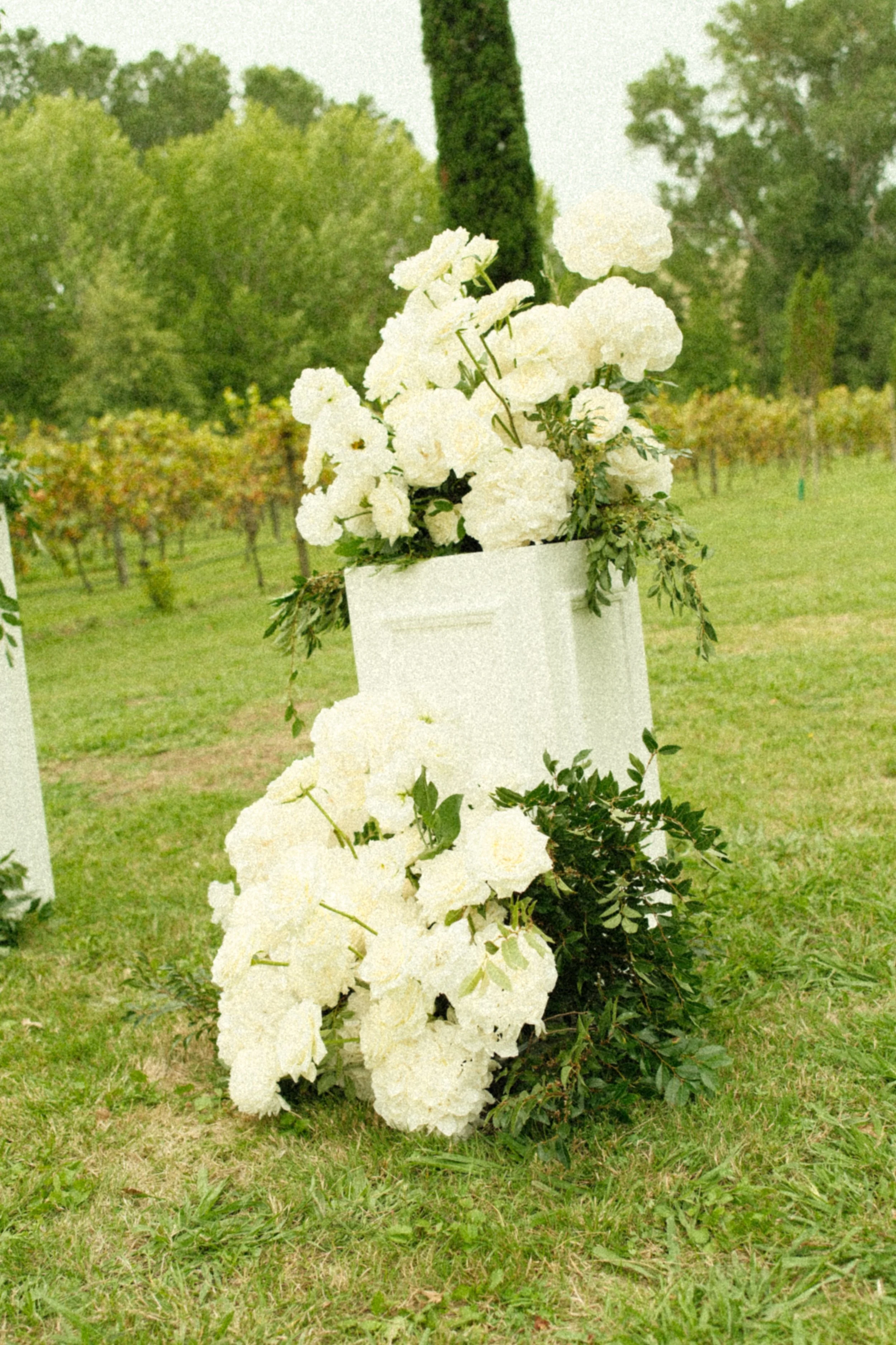 White floral arrangement with hydrangeas and greenery, placed on a white stand outdoors in a lush green park or garden.