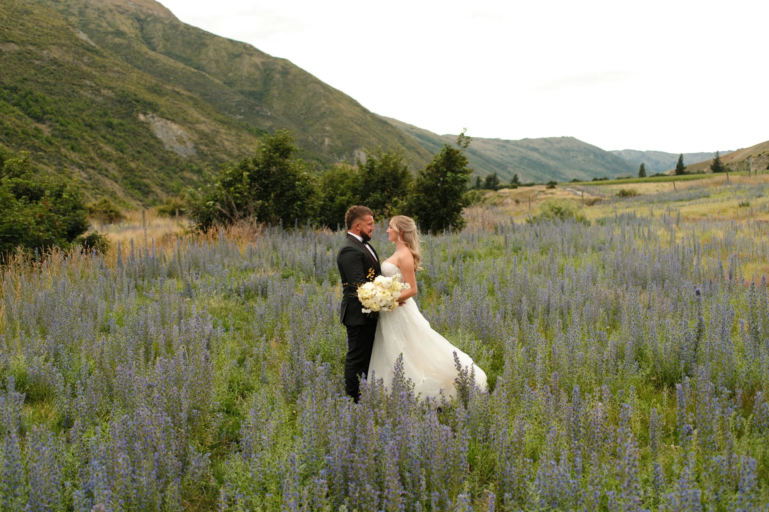 A bride and groom standing in a lavender field with green hills in the background, holding a bouquet of flowers and gazing at each other.