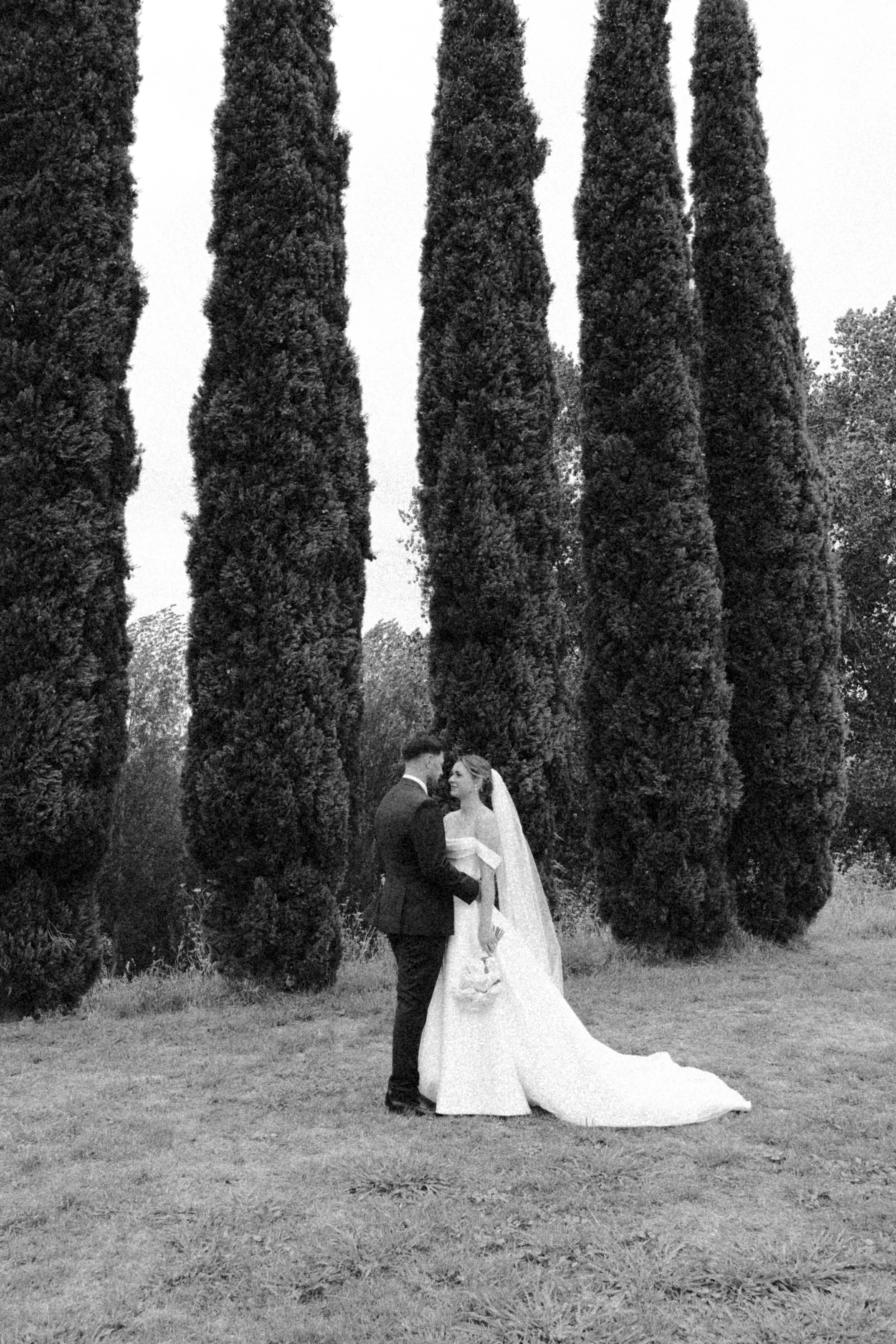 A black-and-white photo of a bride and groom standing close together outdoors, surrounded by tall, narrow trees. The bride is in a wedding gown with a veil, and the groom is in a suit. They appear happy and are holding hands.