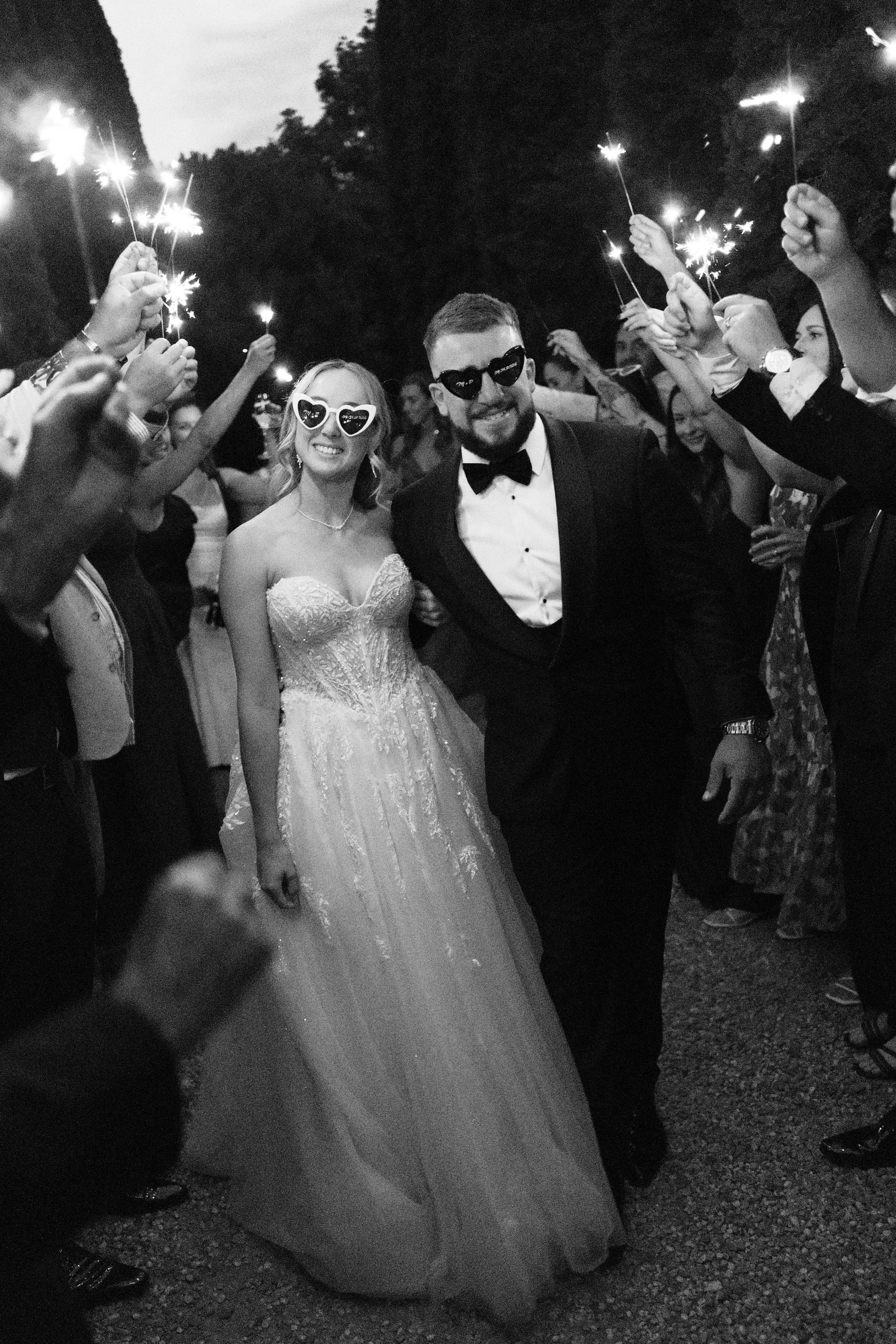 A black and white photo of a bride and groom smiling and walking through a crowd holding sparklers at night.