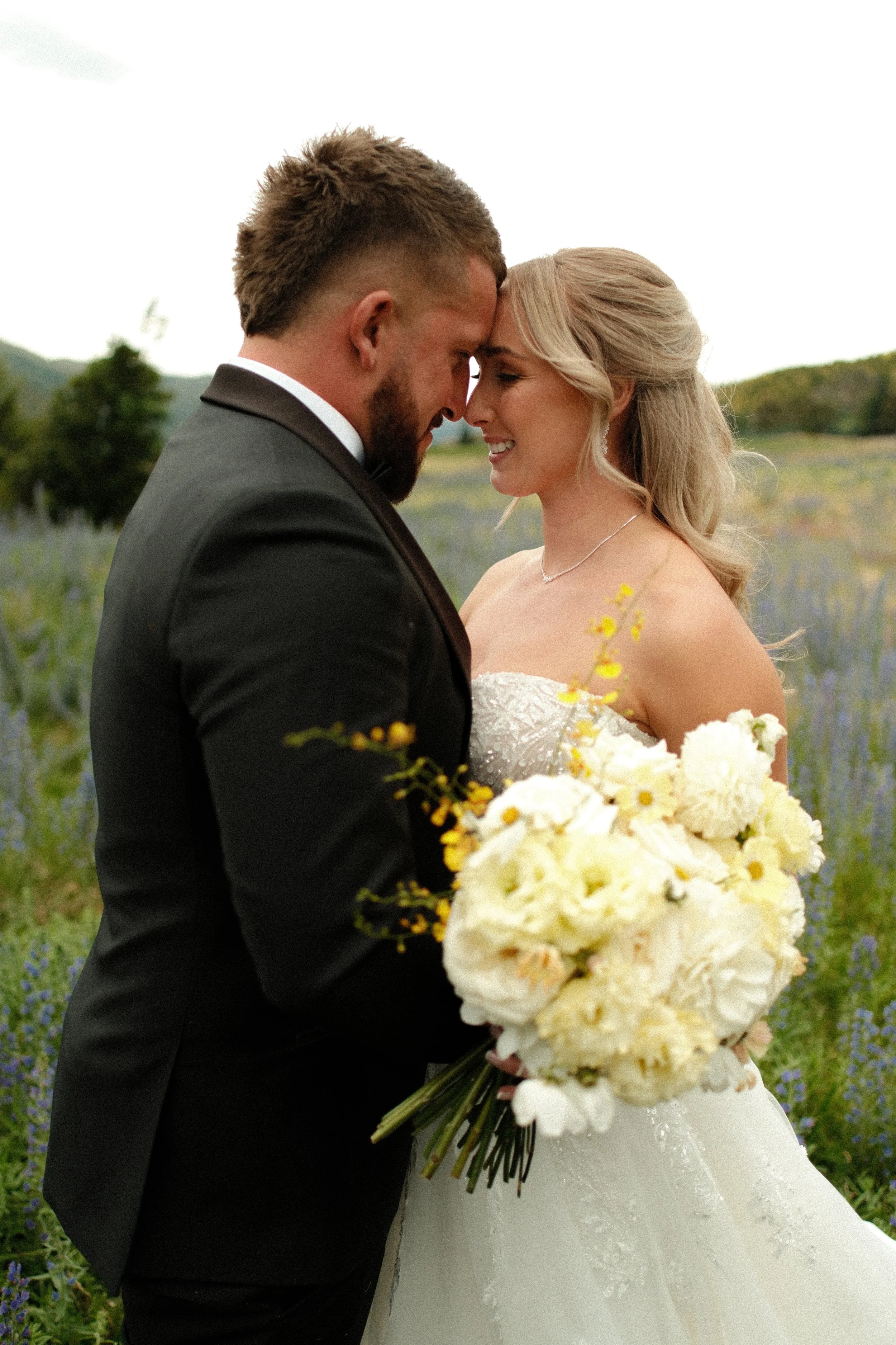 A bride and groom with foreheads touching, holding a bouquet of white flowers, standing outdoors in a field of purple flowers, during daytime.