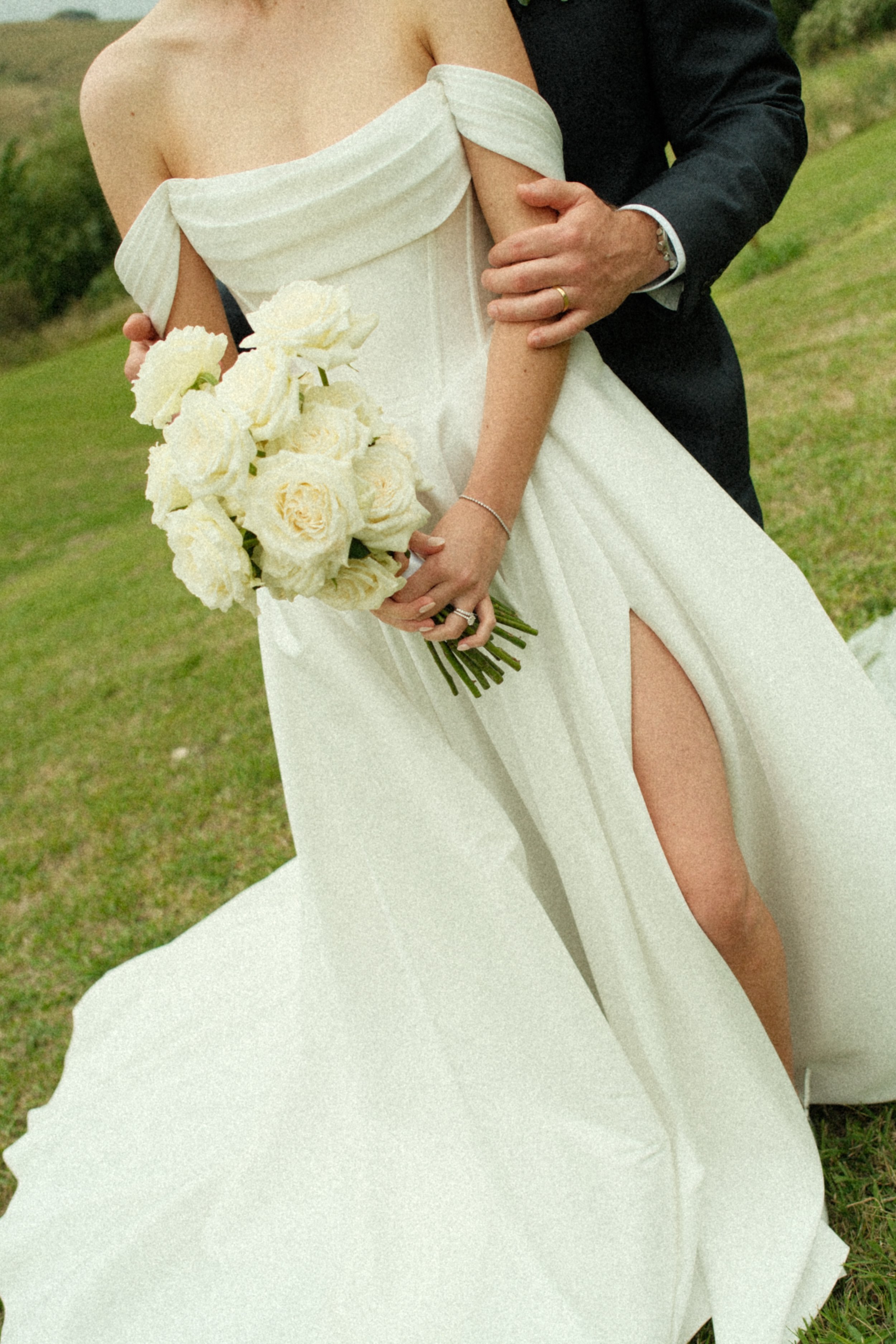 Bride holding a bouquet of white roses, wearing an off-the-shoulder wedding dress with a high slit, standing outdoors with a groom in a dark suit.