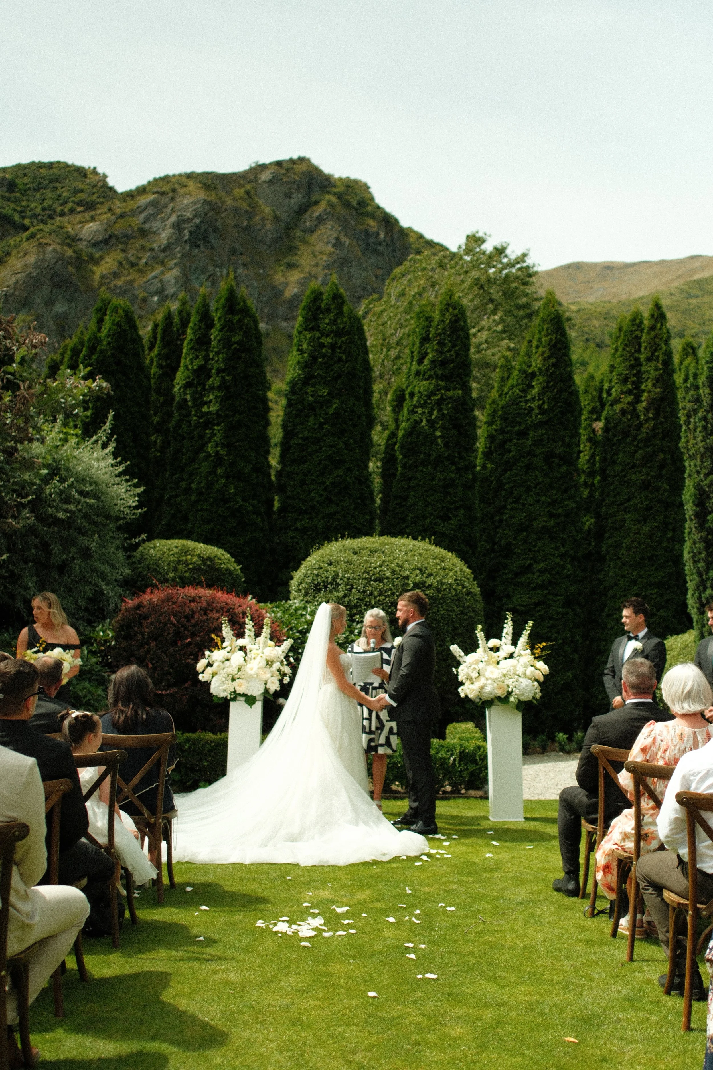 Bride and groom exchanging vows outdoors at a wedding ceremony with guests seated on grass, surrounded by tall green trees and mountains in the background.