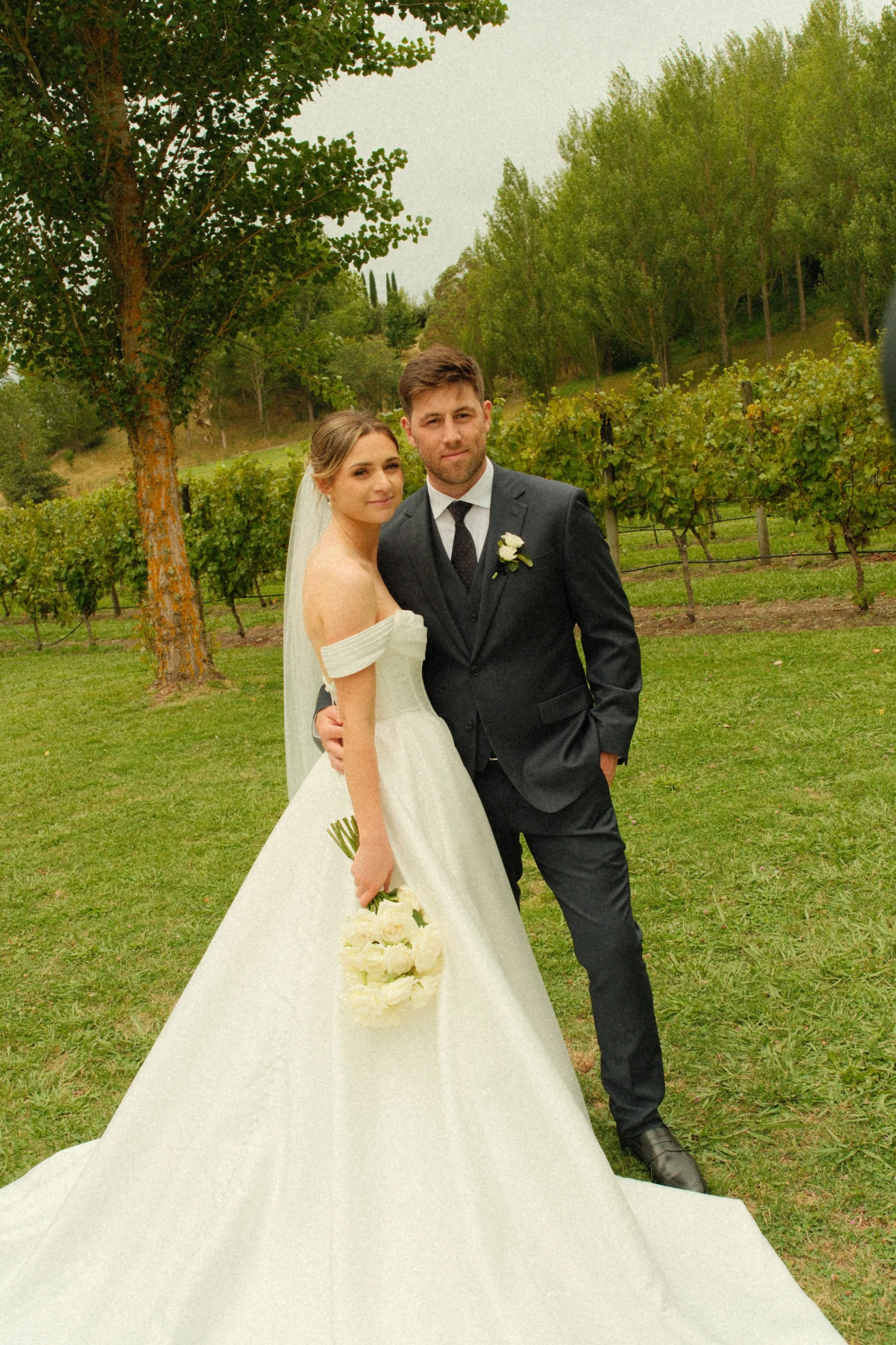 A bride and groom standing together outdoors on their wedding day, with the bride holding a bouquet of white flowers and wearing a white off-the-shoulder dress, and the groom in a dark suit and tie, surrounded by green trees and vineyard.