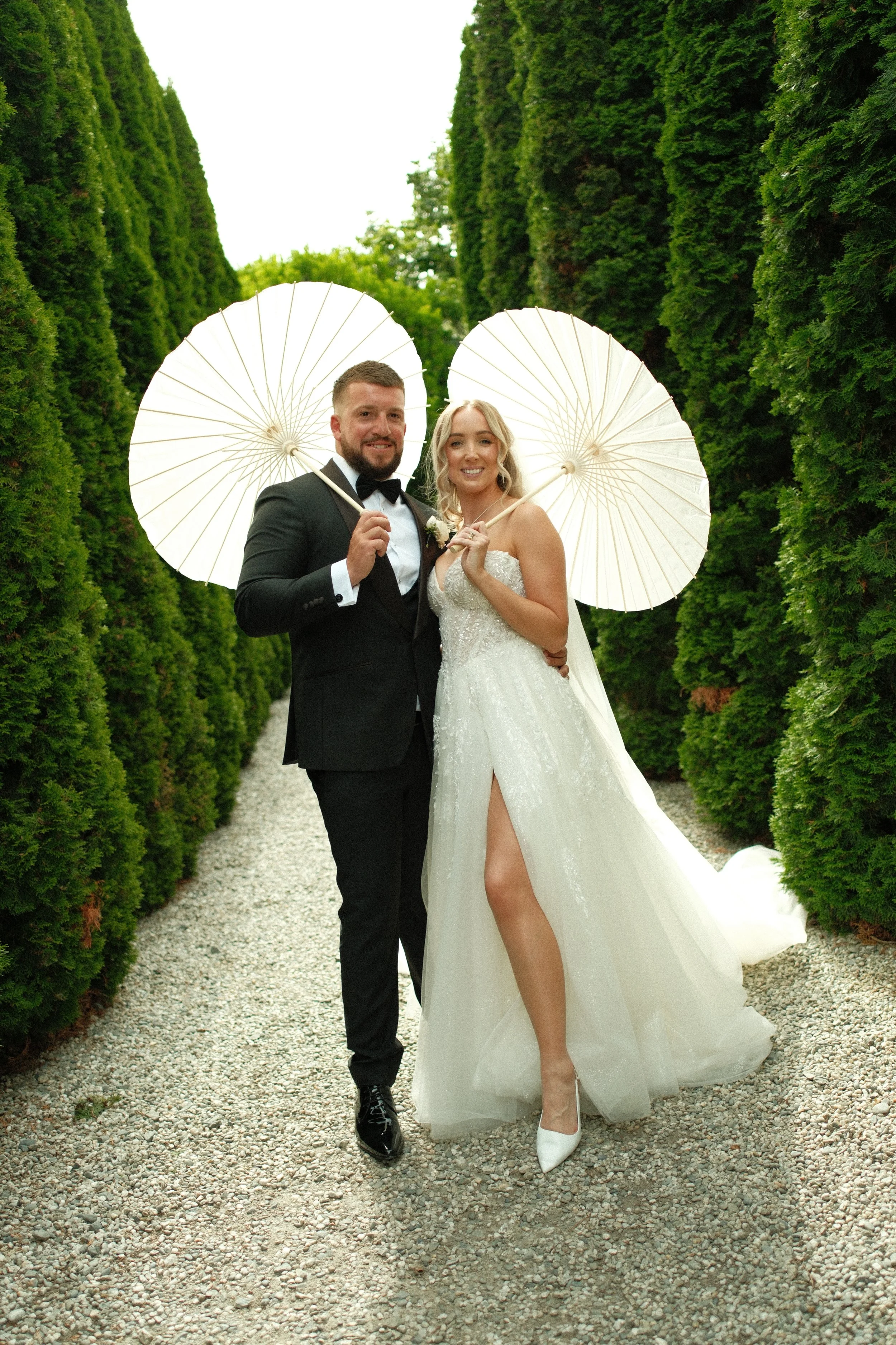 A newlywed couple in wedding attire standing on a gravel path, holding white parasols, surrounded by tall, lush green hedges.