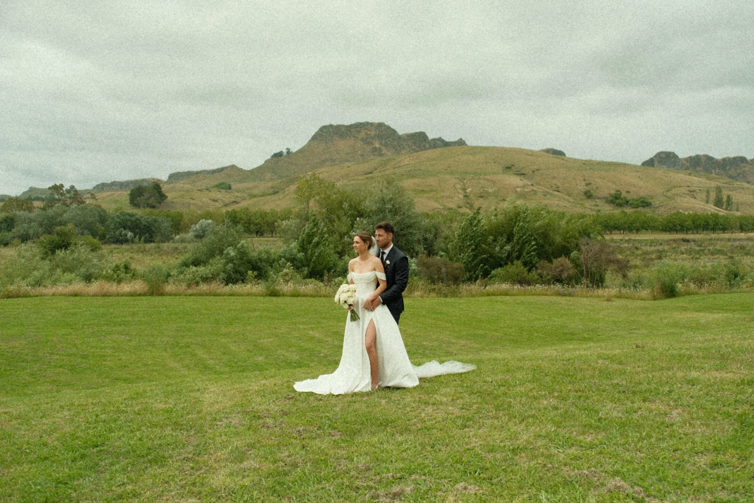 Bride and groom standing on a grassy field with a mountain backdrop, bride holding a bouquet of white flowers, bride in a white wedding dress with a slit, groom in a dark suit, overcast sky.