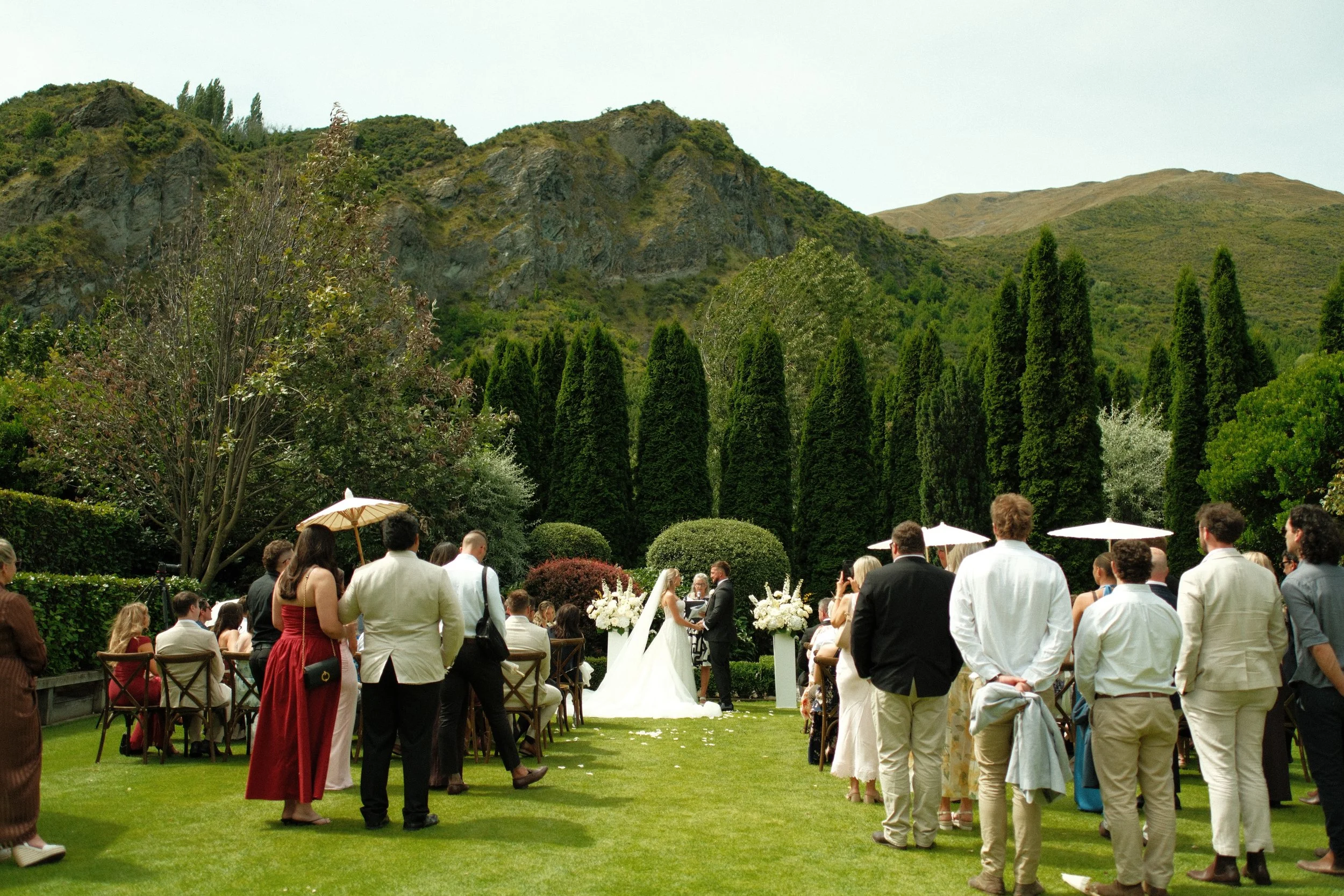 Outdoor wedding ceremony with guests standing and seated, officiants and bride and groom in front, surrounded by lush greenery and mountains.
