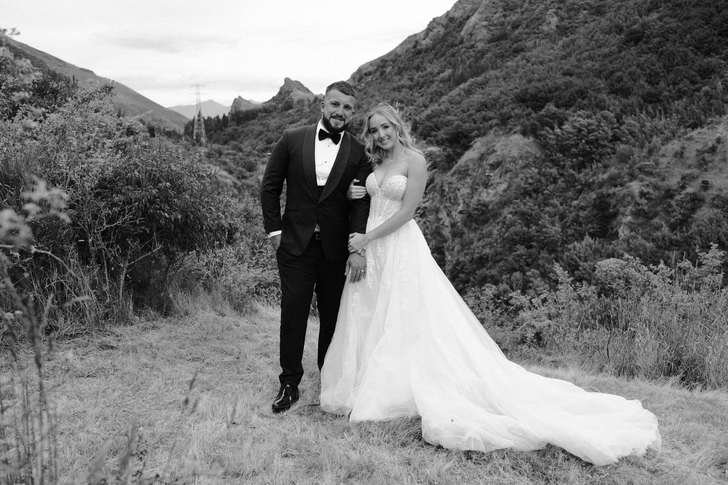 A bride and groom standing outdoors on a grassy area with mountains and trees in the background, dressed in wedding attire, smiling and posing for a photo.