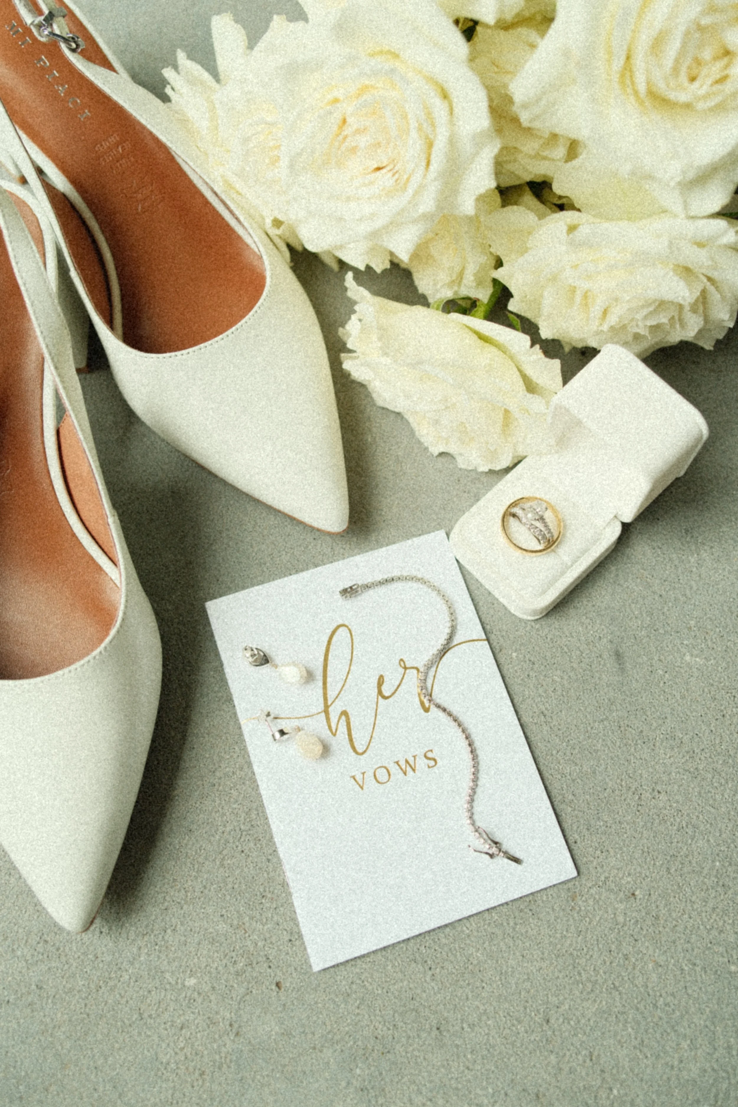 A bridal jewelry set with earrings and necklace, a ring in a box, white high-heeled shoes, and white roses on a surface.