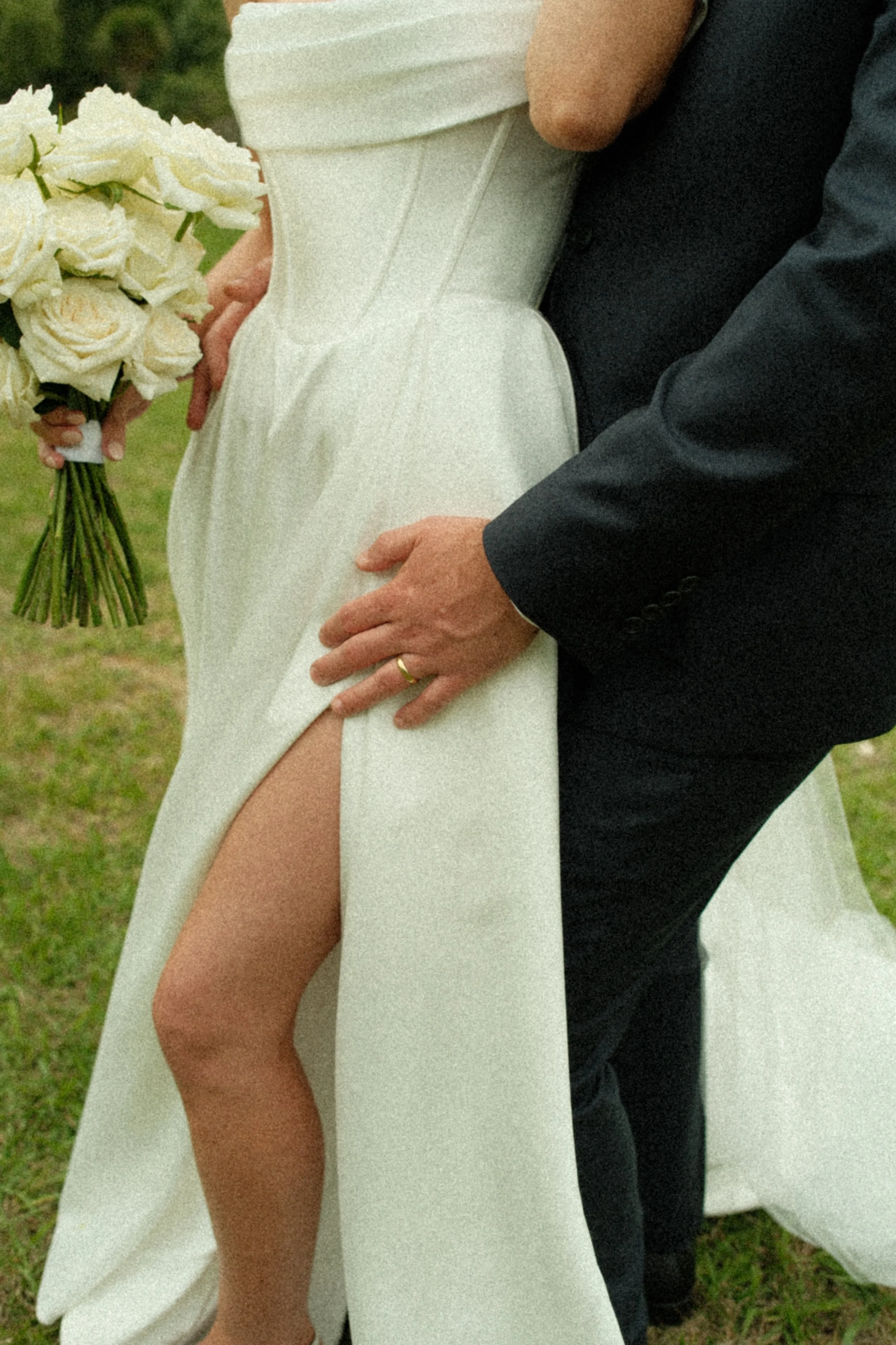 Close-up of a bride and groom at a wedding, with the groom holding the bride's leg to reveal her high slit dress and bouquet, outdoors on grass.