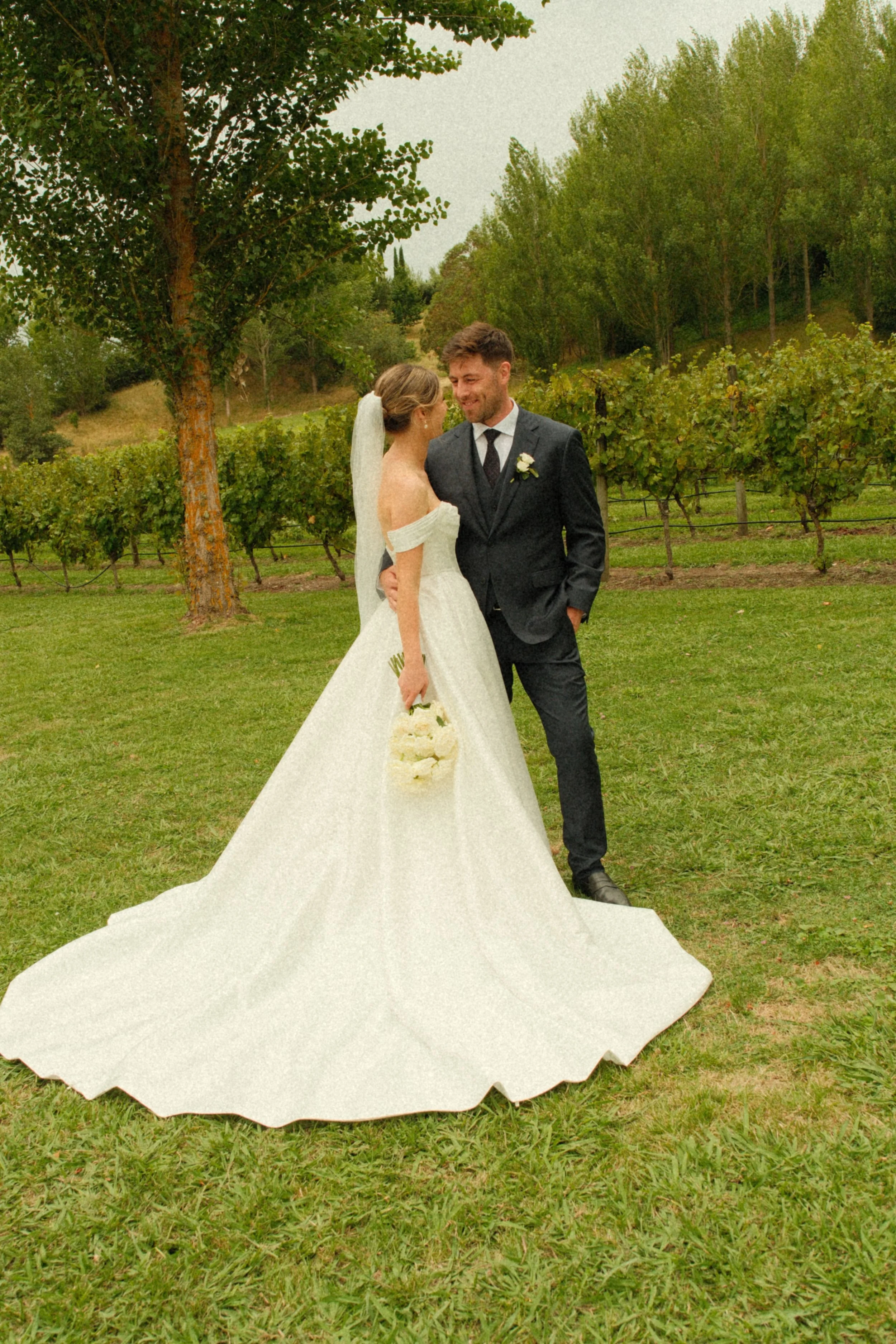 A bride and groom standing close together on a grassy field during her wedding, with the bride holding a bouquet of white flowers and a vineyard in the background.