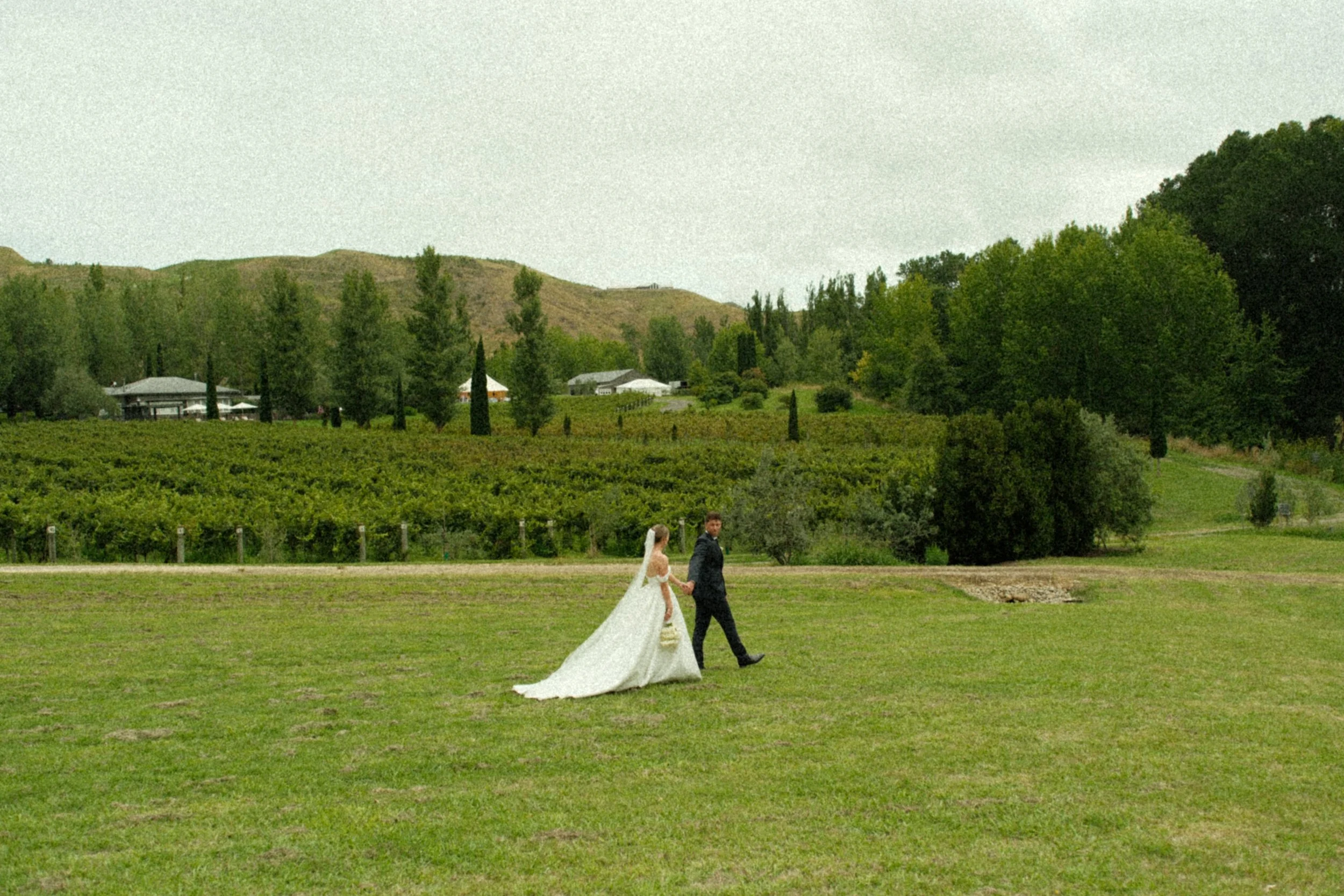 A bride and groom walking hand in hand on a lush green field with trees and hills in the background.