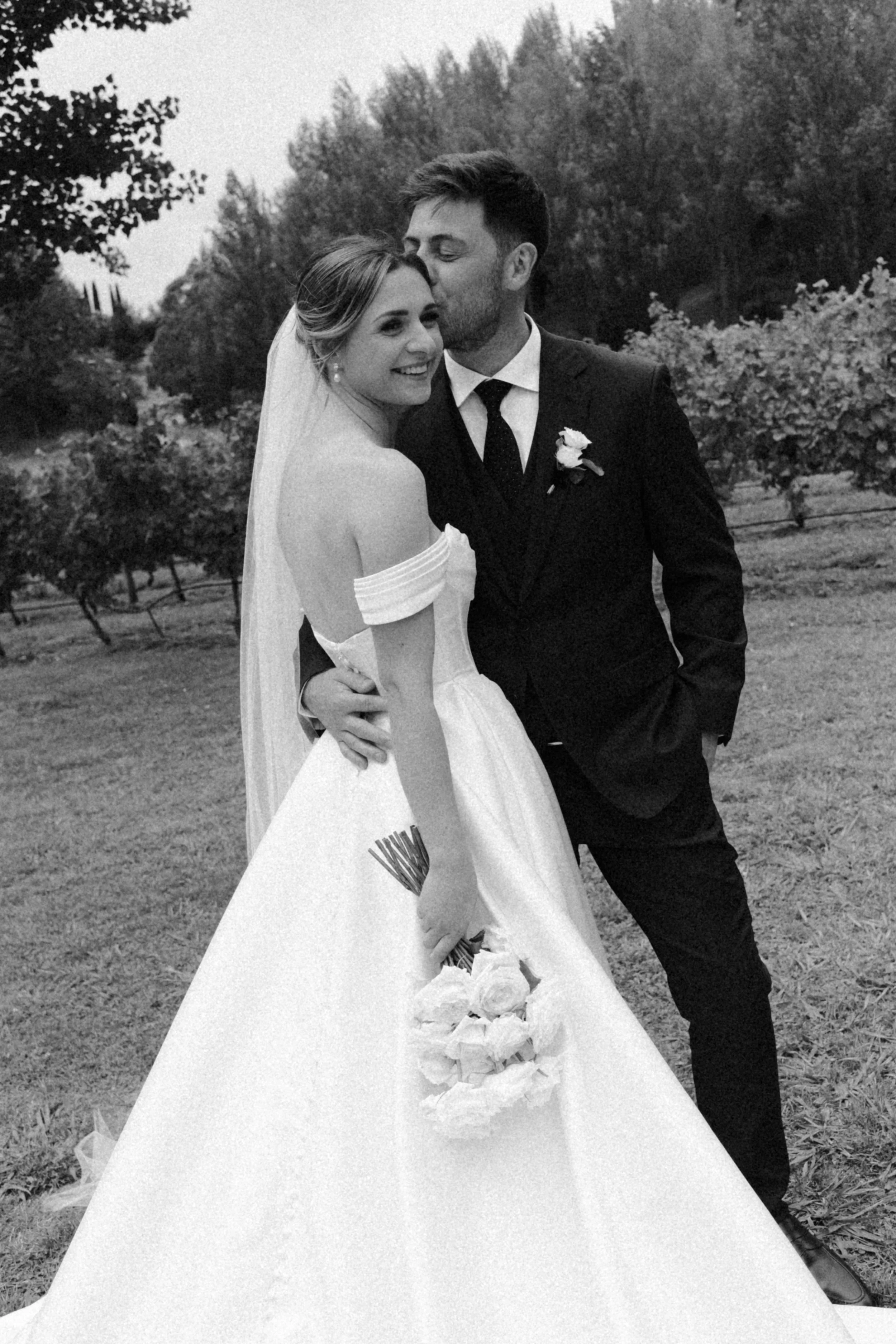Black and white photo of a bride and groom standing outside, smiling and hugging. The bride is holding a bouquet of roses, and the groom is kissing her head.