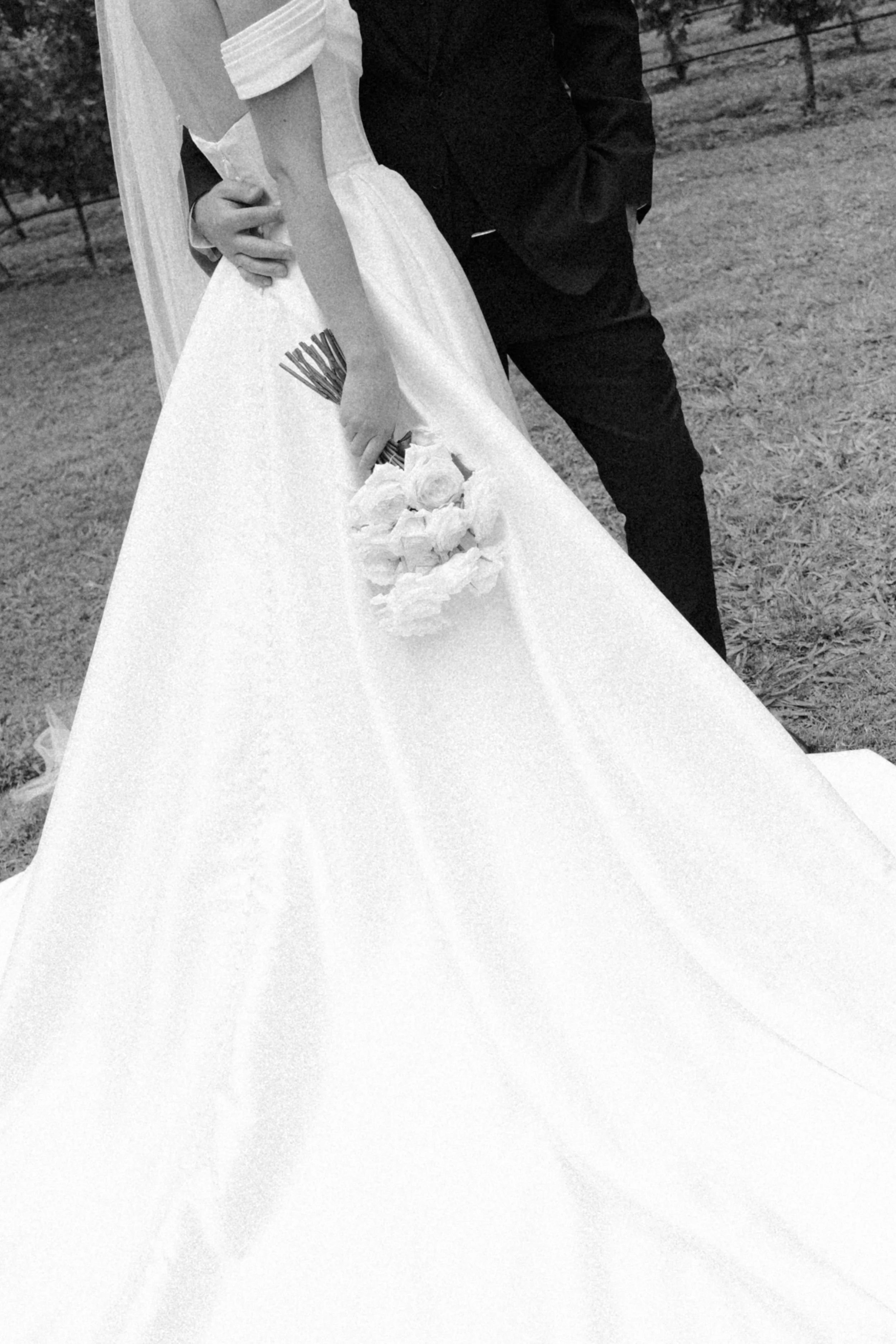 Black and white photo of a bride and groom, with the bride holding a bouquet of flowers, standing outdoors on grass.