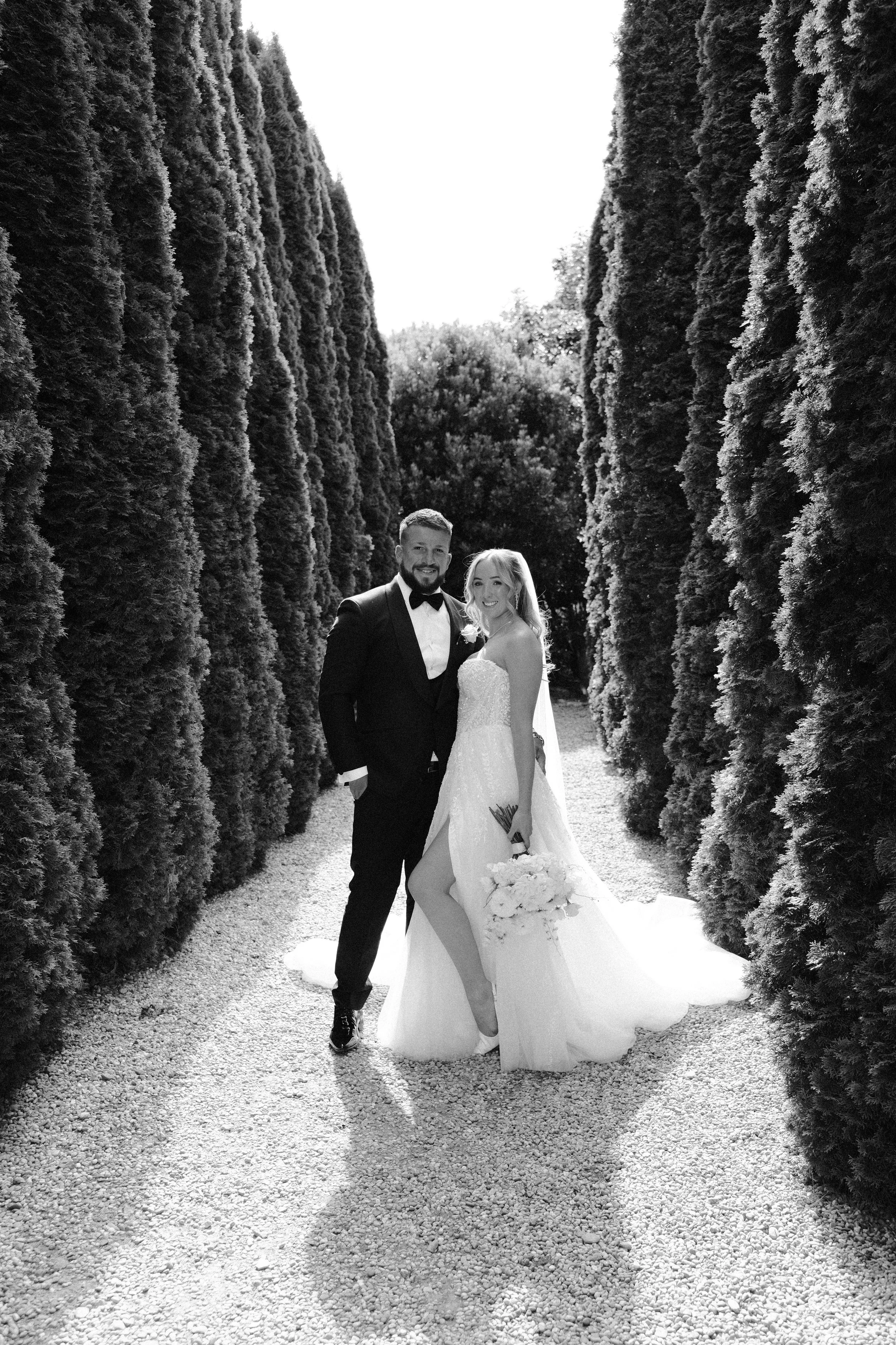 Black and white photograph of a bride and groom standing together outdoors between tall, narrow trees. The bride is holding a bouquet and wearing a strapless wedding dress with a high slit, while the groom is dressed in a tuxedo with a bow tie. They 