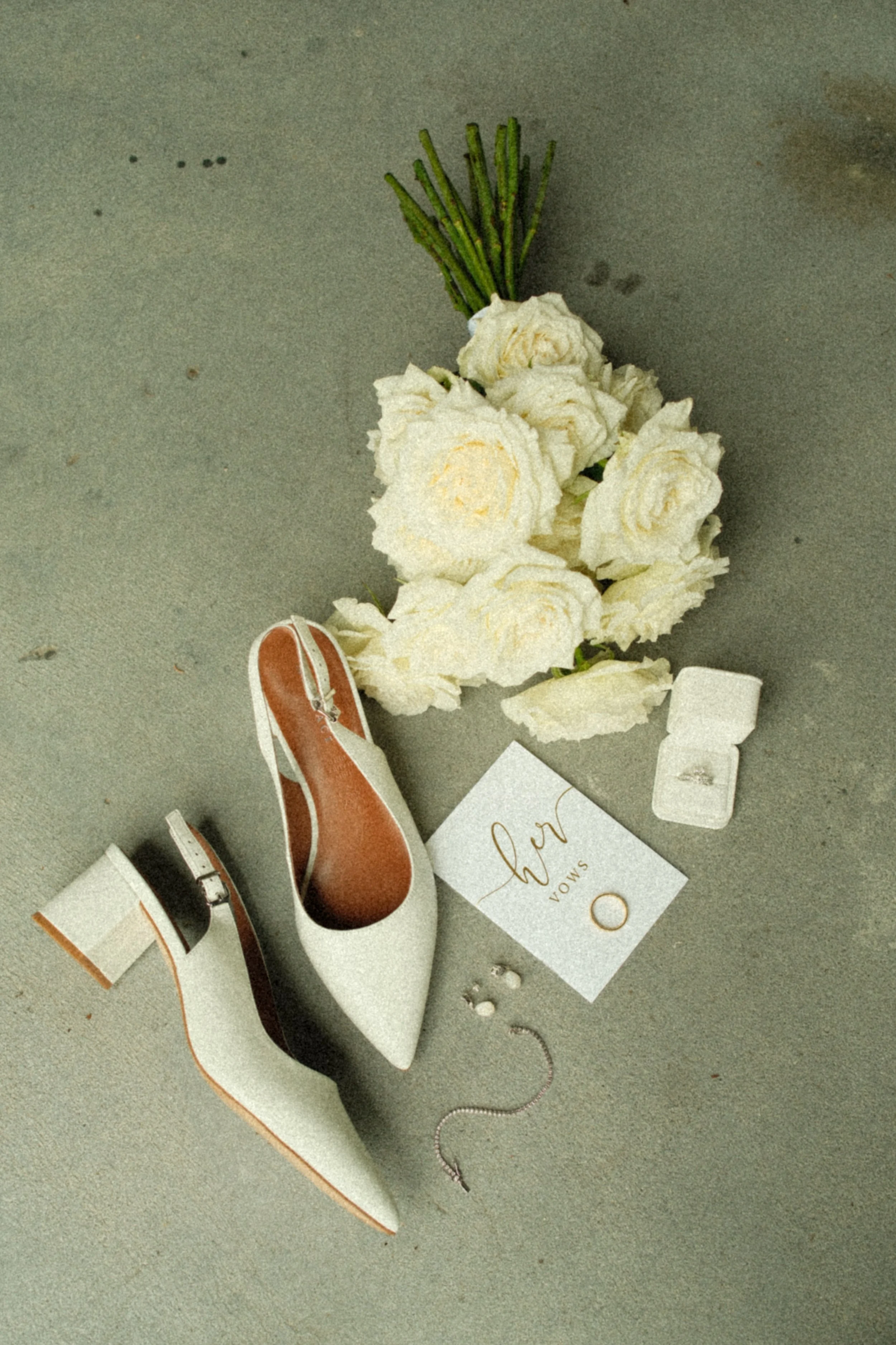 Wedding accessories including a white high heel shoe, a bouquet of white roses, a vow card, a small gift box, and jewelry laid out on a concrete floor.