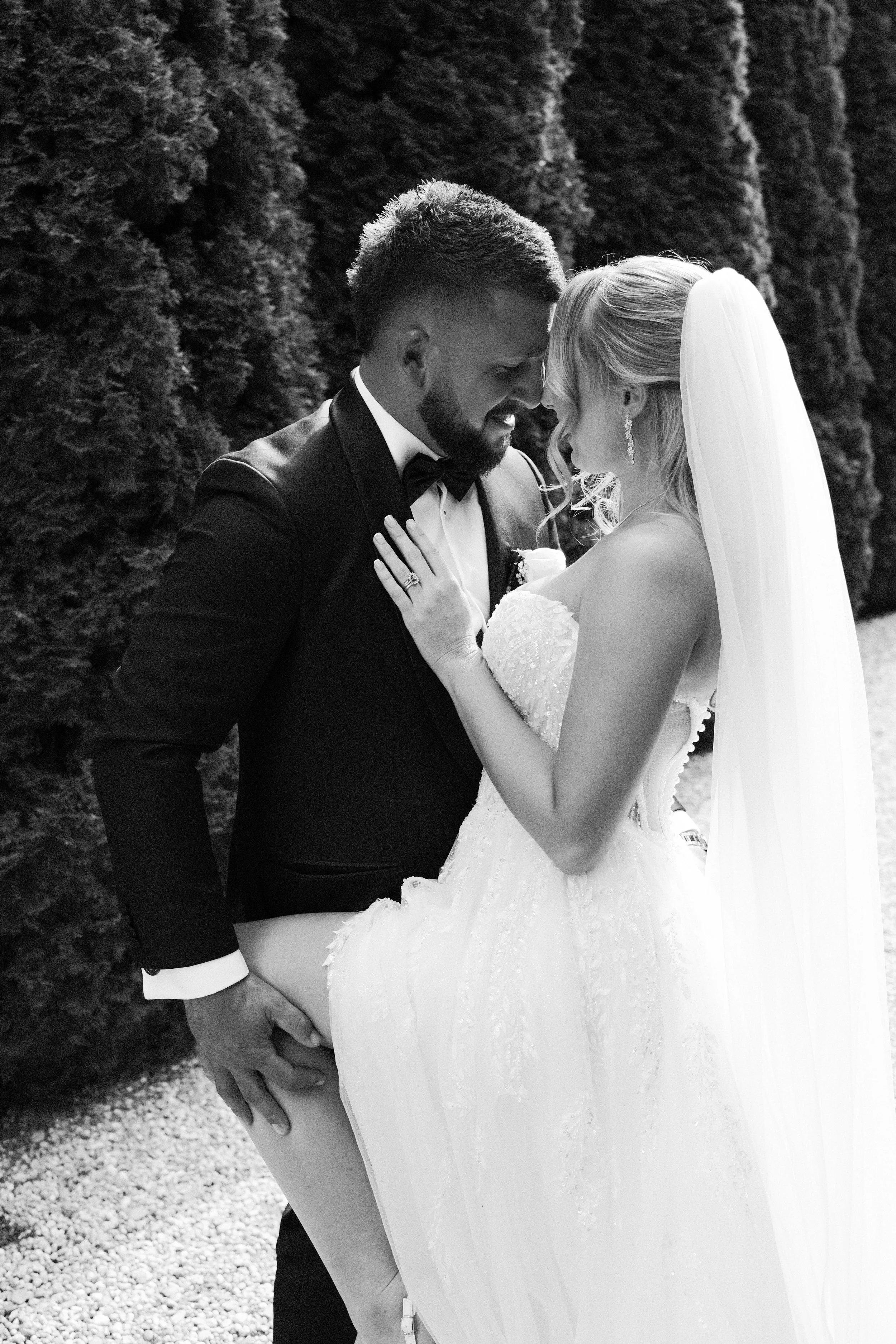 Black and white photo of a bride and groom on their wedding day, with the groom lifting the bride, who is smiling and wearing a strapless wedding gown with a long veil, in front of tall trees.
