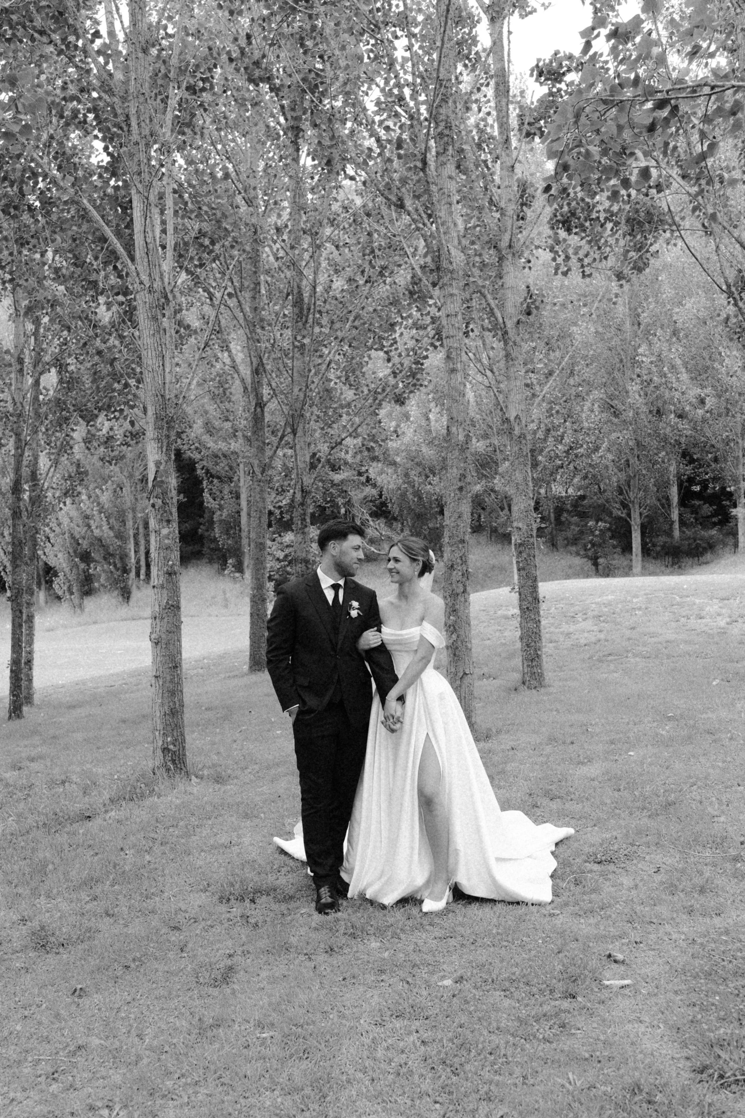 A black-and-white photograph of a bride and groom holding hands and walking together outdoors among trees during daytime.