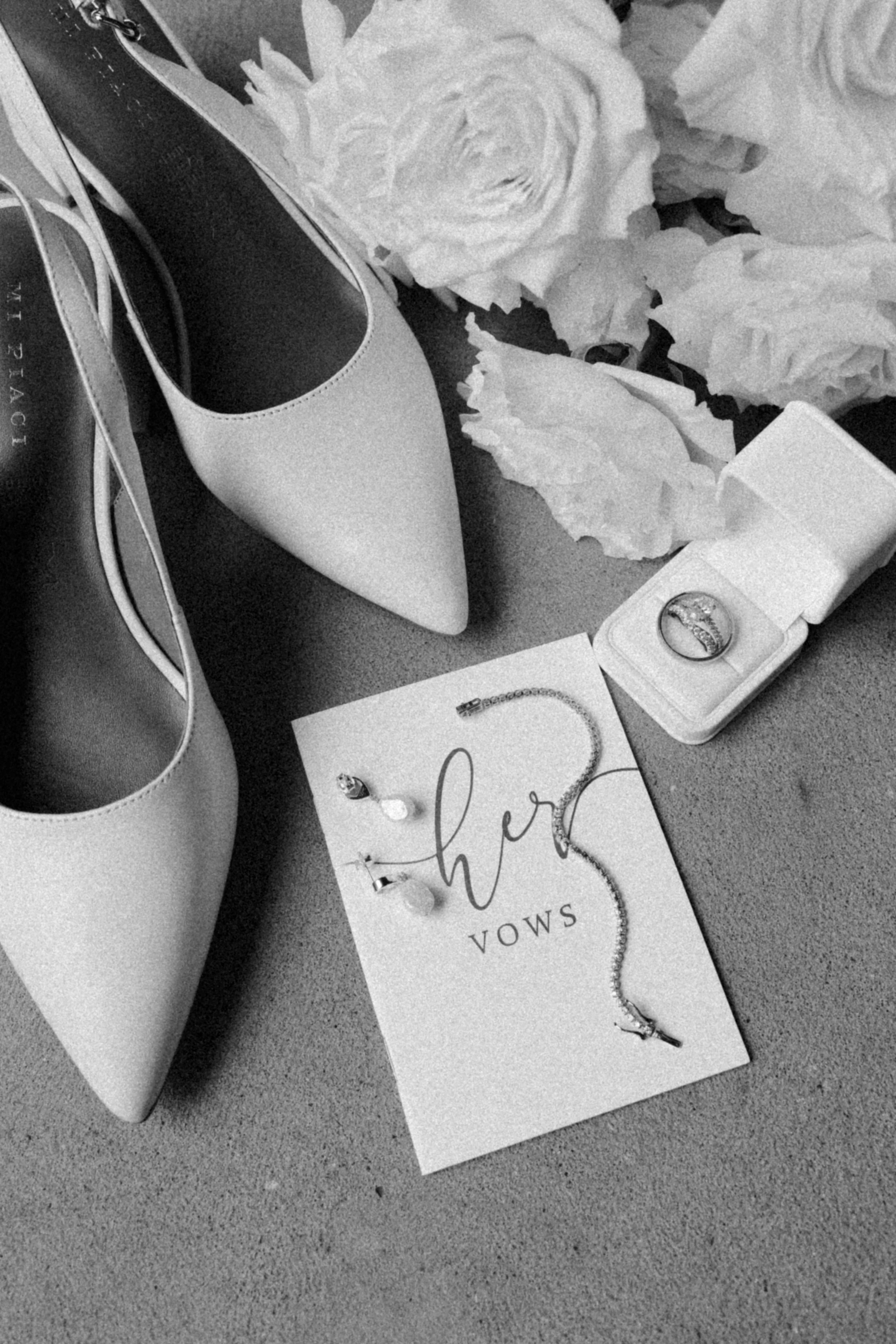A pair of light-colored high heels, a jewelry box with a ring, a necklace and earrings on a card with the word 'her' and 'VOWS,' white flowers, and a love card with rings on it.