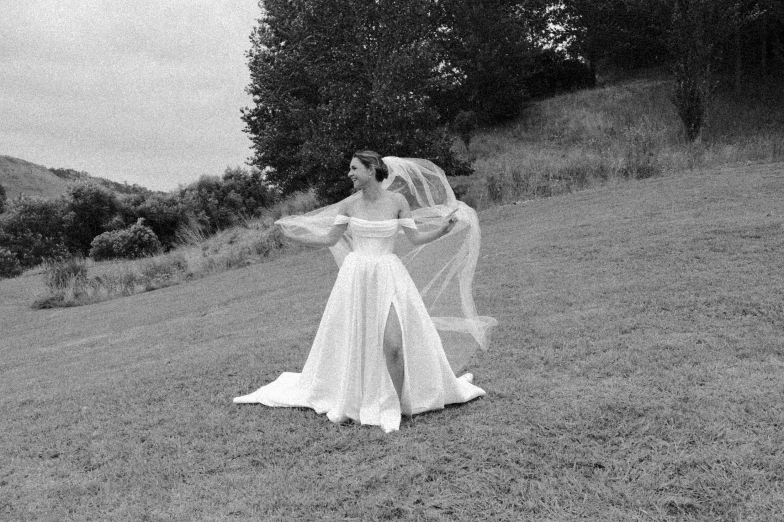 A smiling woman in a vintage wedding dress standing on a grassy field holding a sheer veil behind her in a rural outdoor setting with trees and hills in the background.