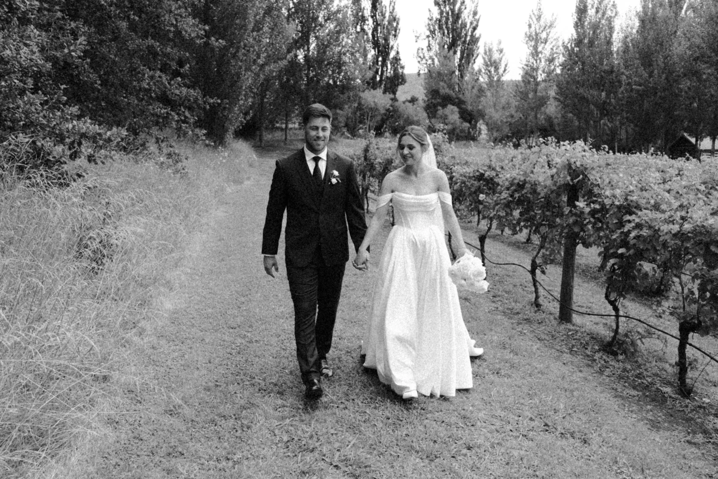 A bride and groom walking hand in hand through a vineyard during their wedding, smiling.