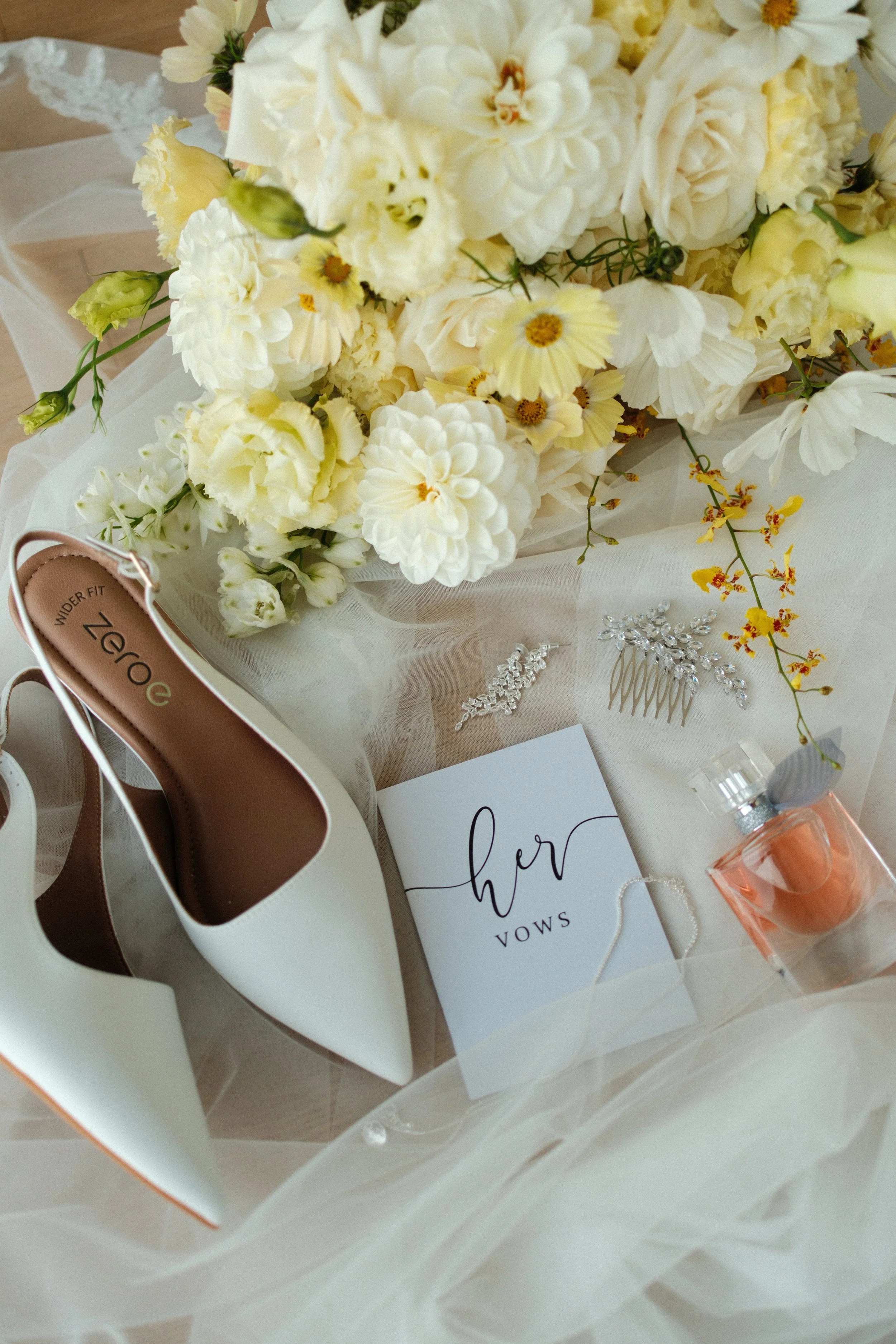 A wedding vow card, white high heels, a bridal bouquet of white flowers, and jewelry and perfume items on a soft fabric surface.