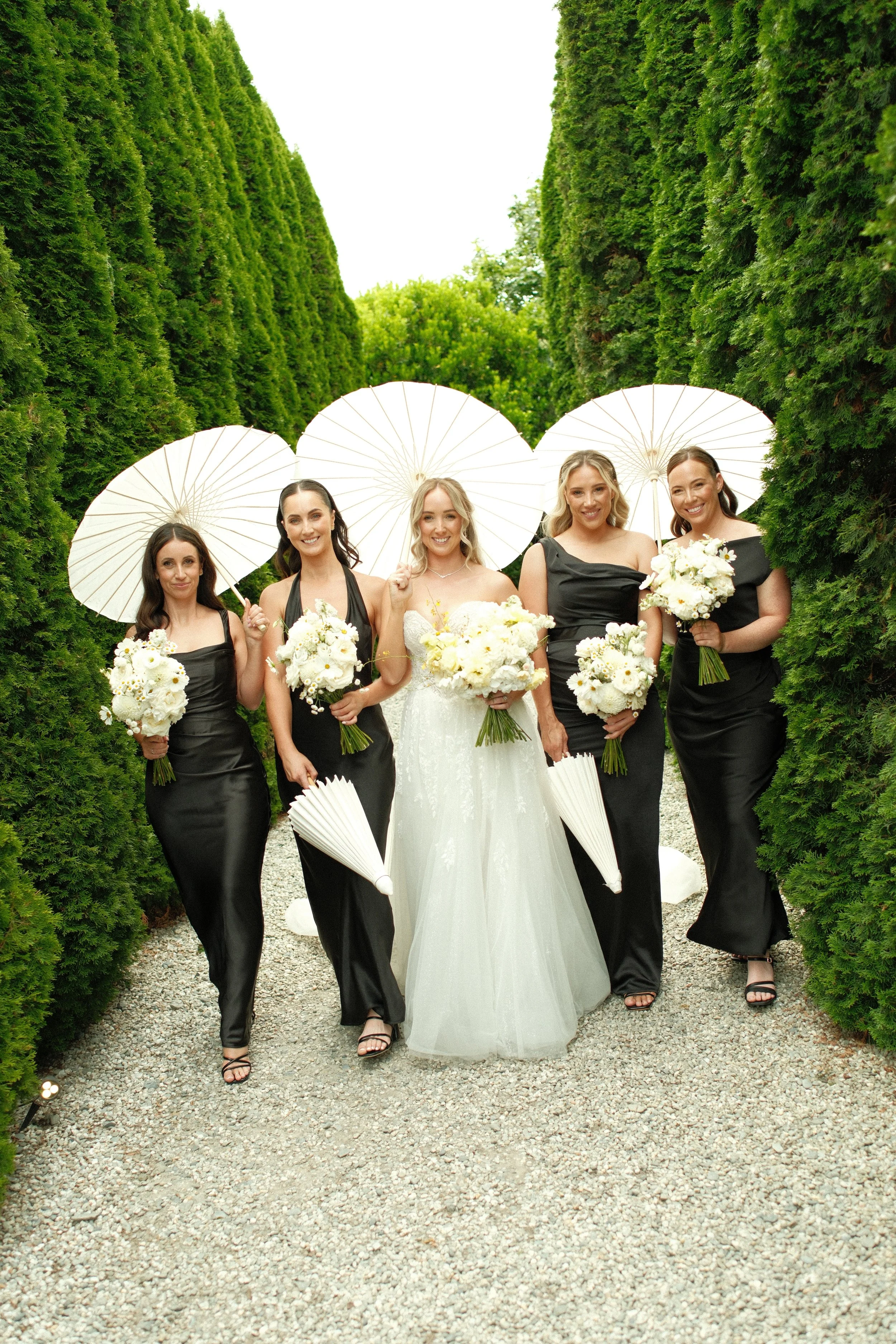 Bridal party walking through greenery pathway, holding bouquets and parasols.