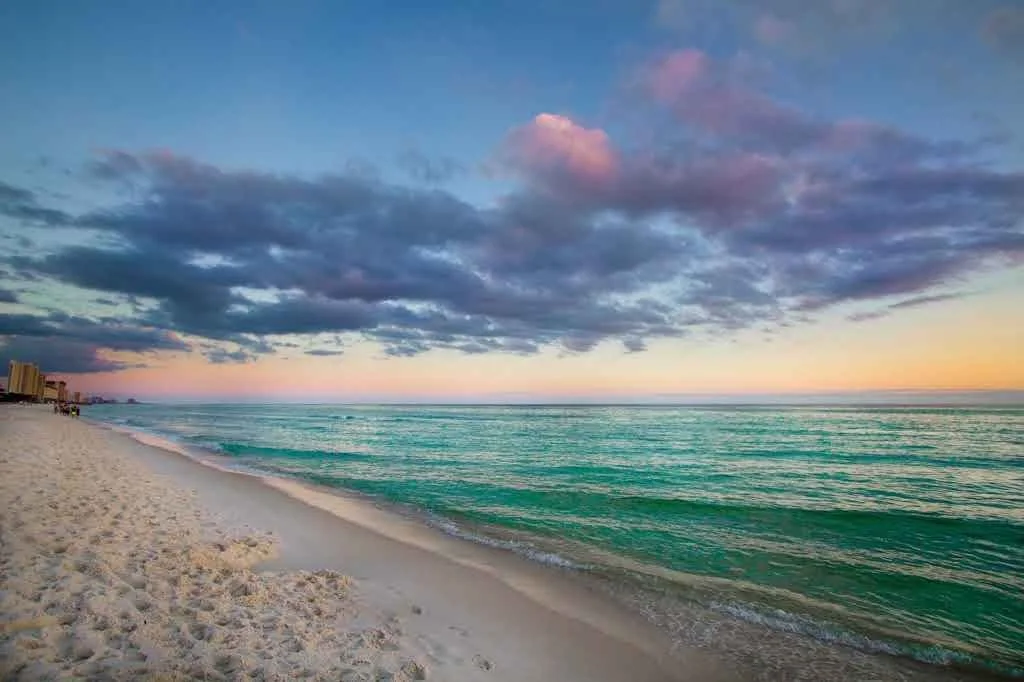 A serene beach scene at sunset with white sand, calm turquoise water, and a sky filled with blue and pink clouds.