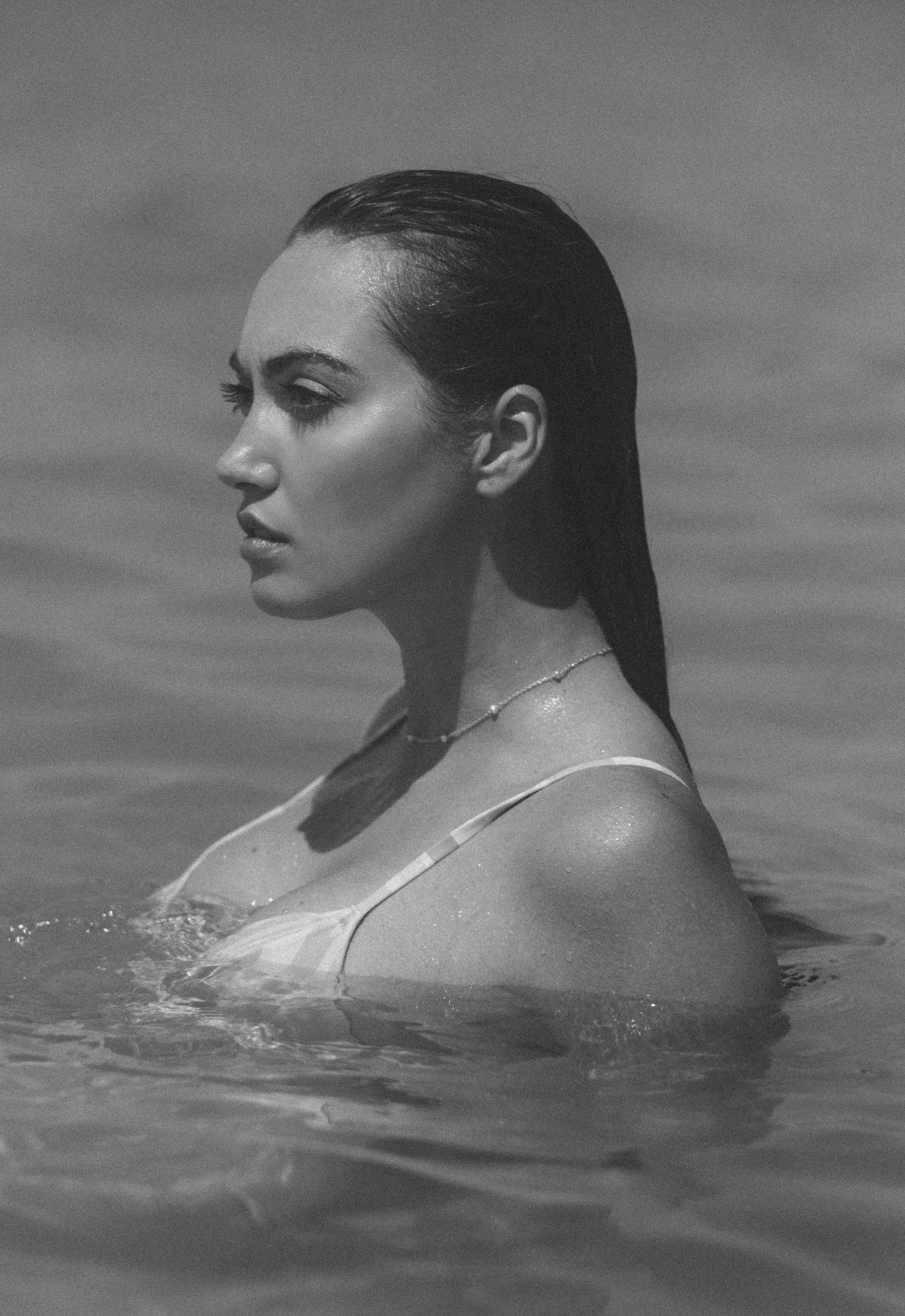 A black-and-white photo of a woman with long, wet hair, in the water, looking to the side with a serene expression, wearing a swimsuit and a necklace.