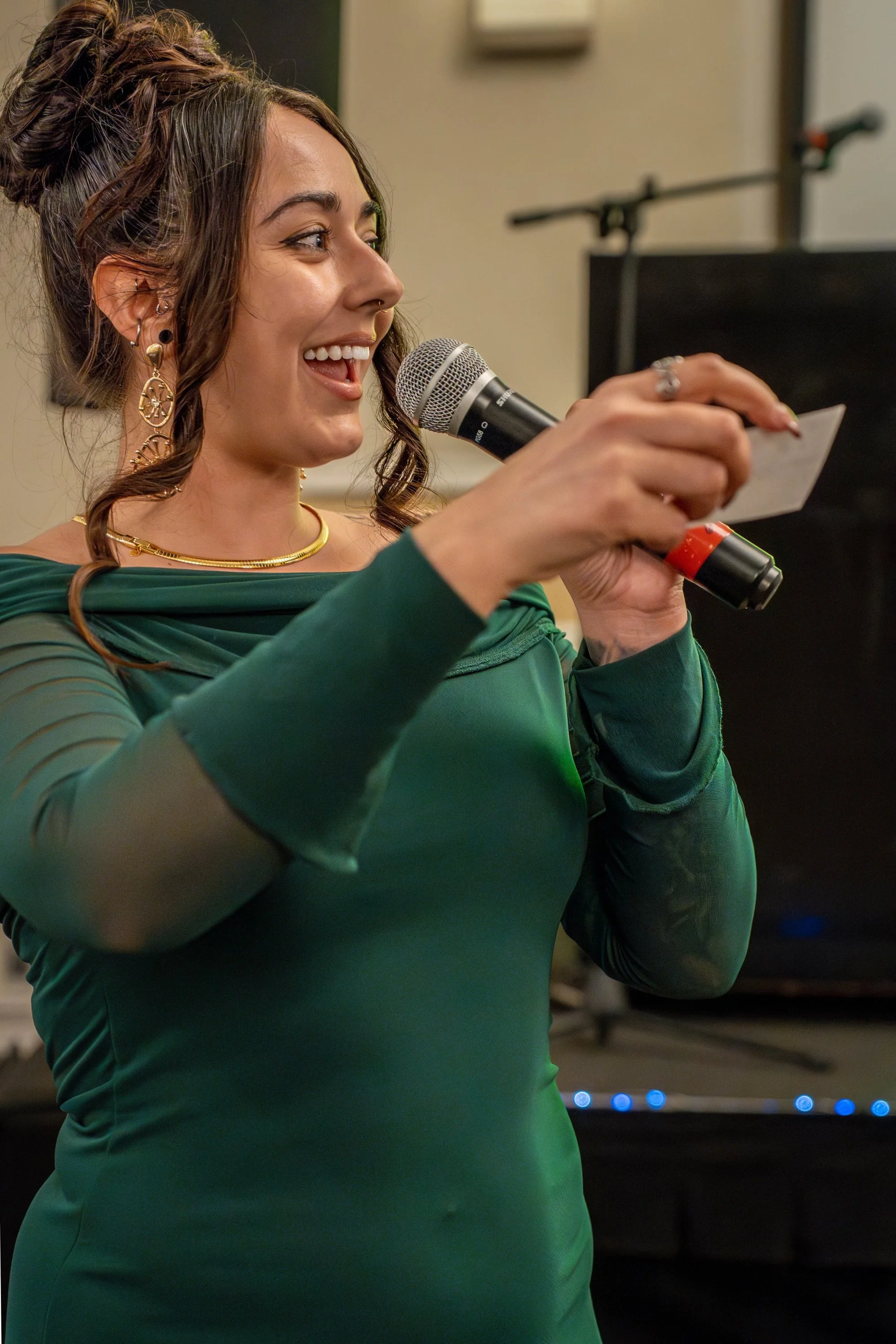 A woman with dark brown hair styled in an updo with loose curls, wearing large gold jewelry including earrings and a necklace, in a green dress, smiling while holding a microphone.
