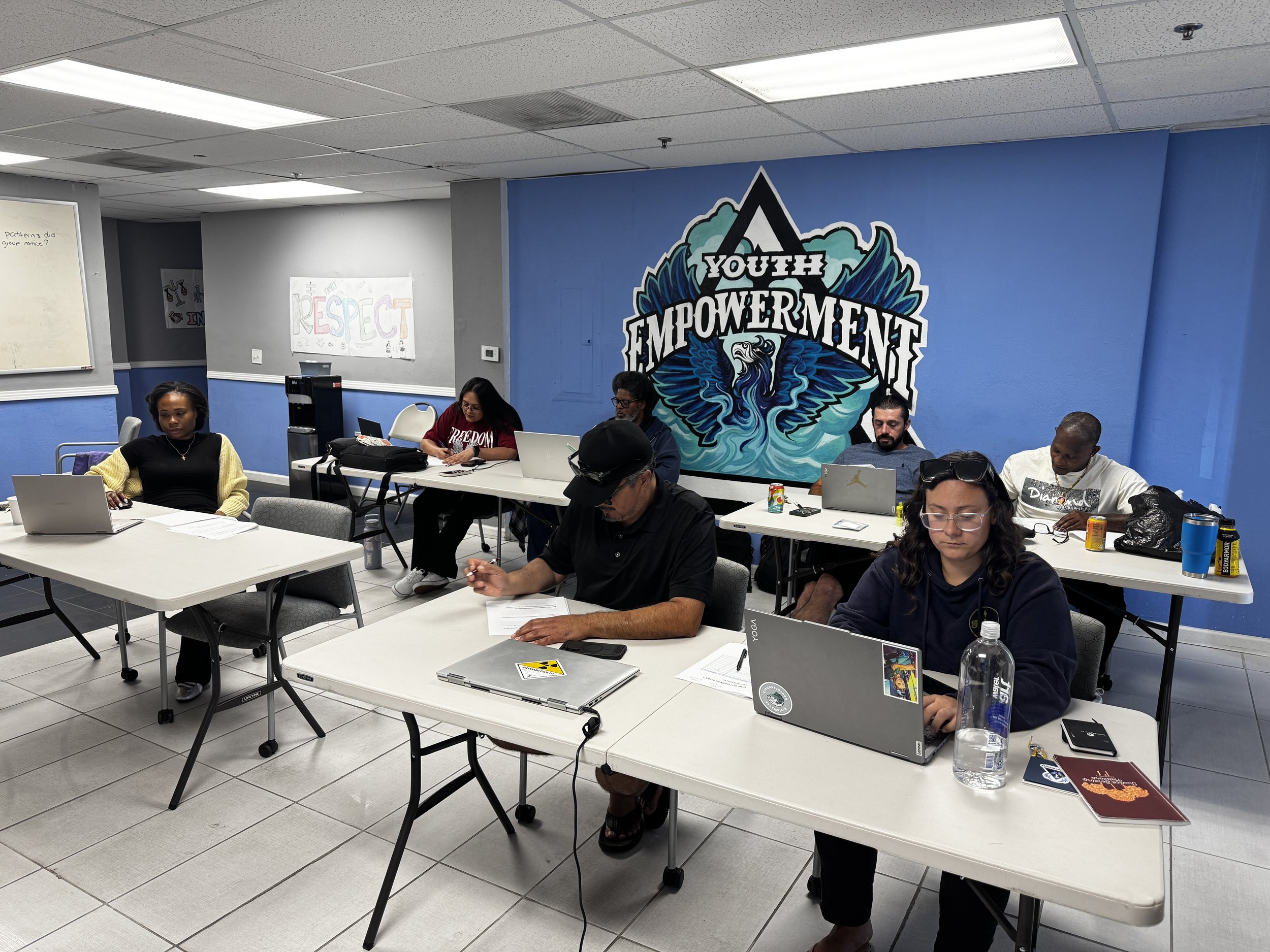 A group of diverse people attending a meeting or workshop in a room with blue walls, a large mural with the words 'Youth Empowerment', and posters on the wall. They are seated at tables with laptops, notebooks, and drinks.
