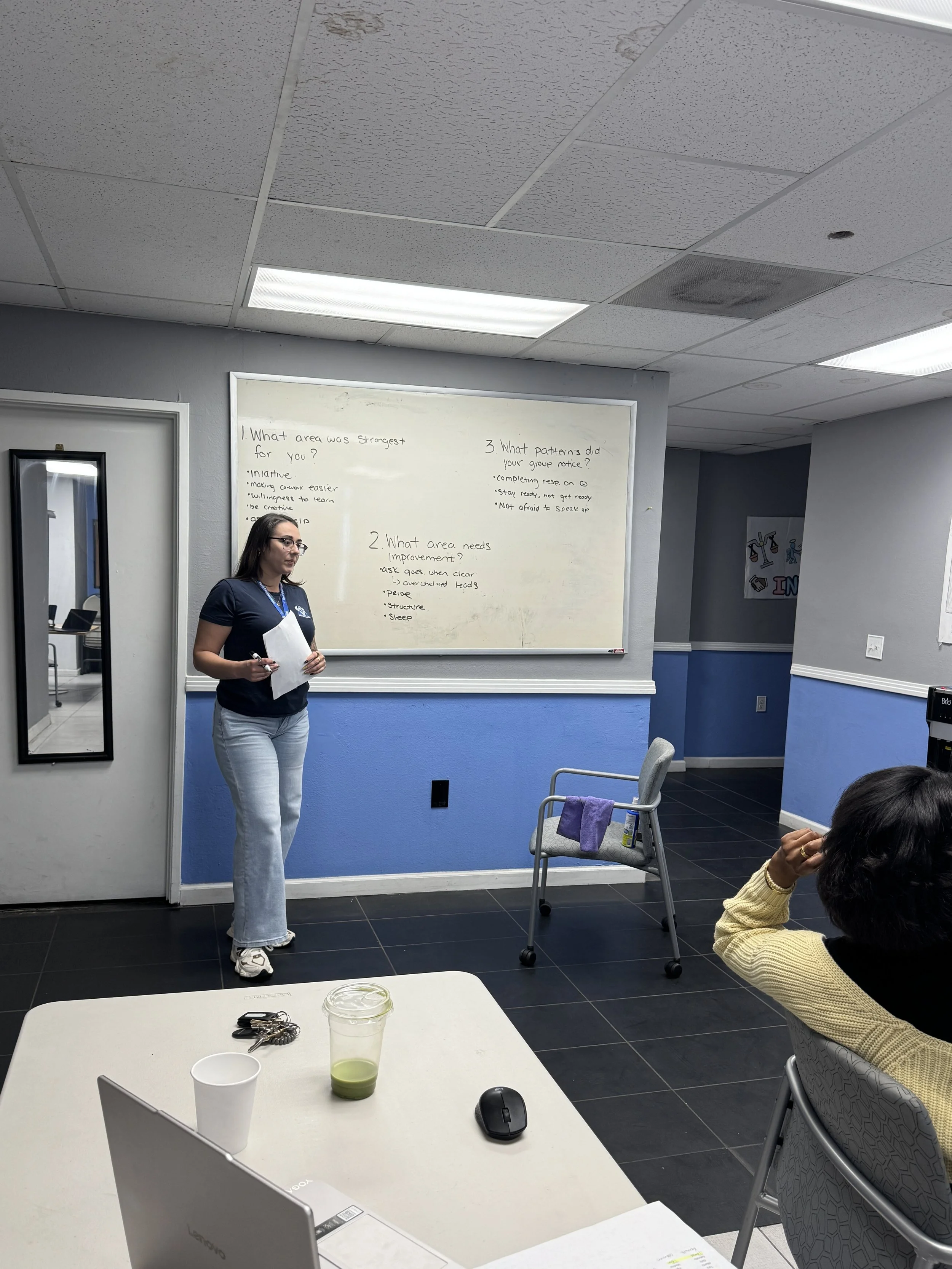 A woman standing in front of a whiteboard giving a presentation in a conference room. The whiteboard has handwritten notes and questions. In the foreground, there is a table with a laptop, a cup, and a drink. A woman seated at the table is partially visible, wearing a yellow sweater.