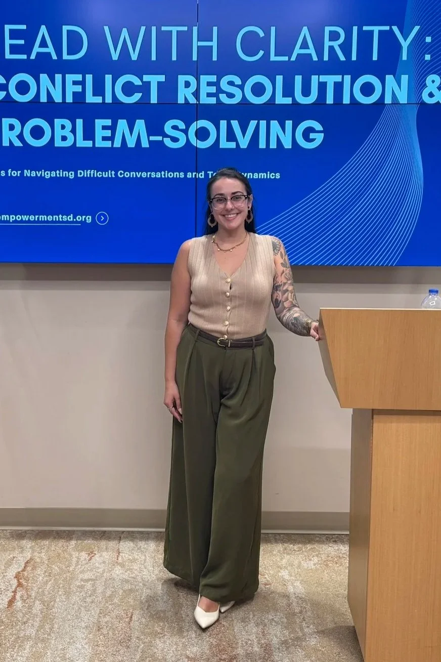 A woman standing next to a podium in front of a large digital display screen with the text "Lead with Clarity: Conflict Resolution & Problem-Solving." She is smiling, wearing glasses, a sleeveless beige vest, high-waisted olive green pants, white pointed shoes, and has a tattoo sleeve on her left arm. She has dark hair, earrings, and a necklace, and is holding the edge of the podium with her right hand.