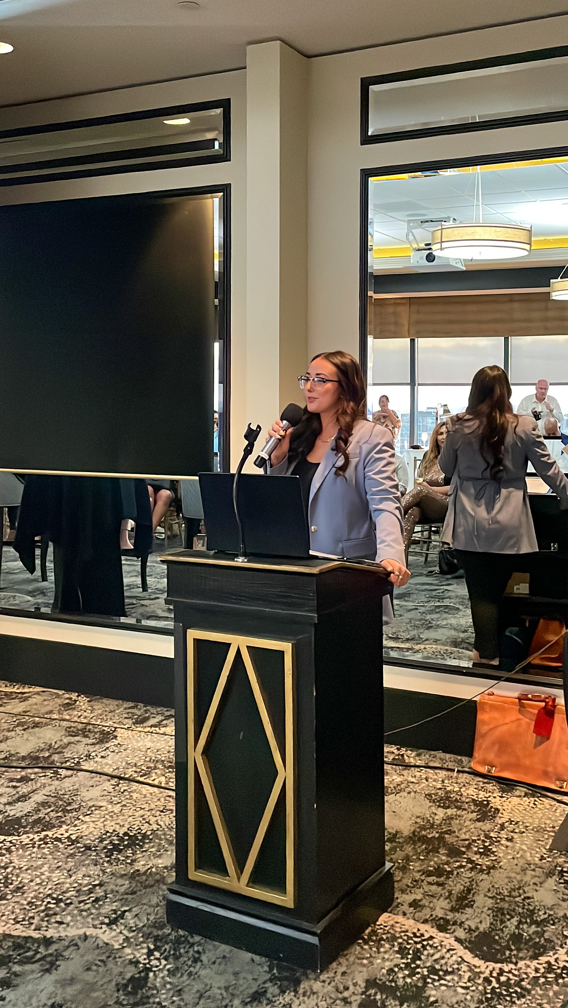 A woman with long brown hair and glasses speaking into a microphone at a podium with a geometric design in a conference room, with people seated and standing in the background.