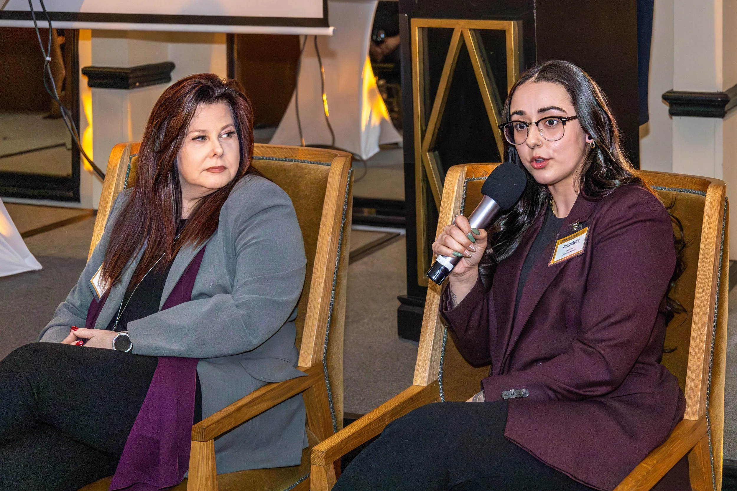 Two women sitting at a table during a discussion or interview in a formal setting, with one woman speaking into a microphone.
