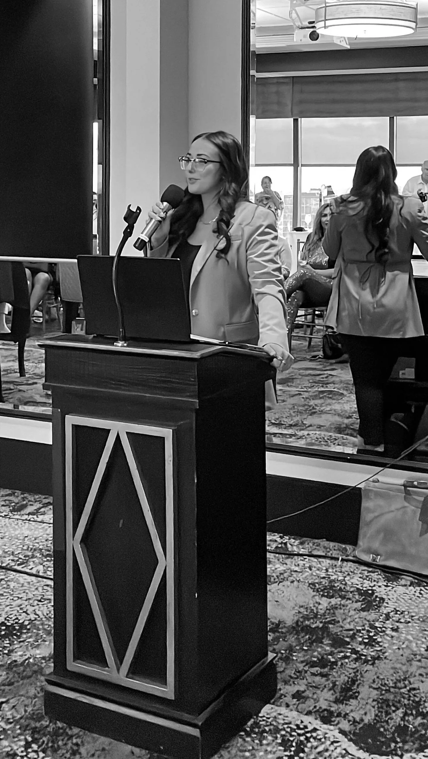 A woman with long hair and glasses speaking into a microphone at a podium during an indoor event, with people seated and standing in the background.