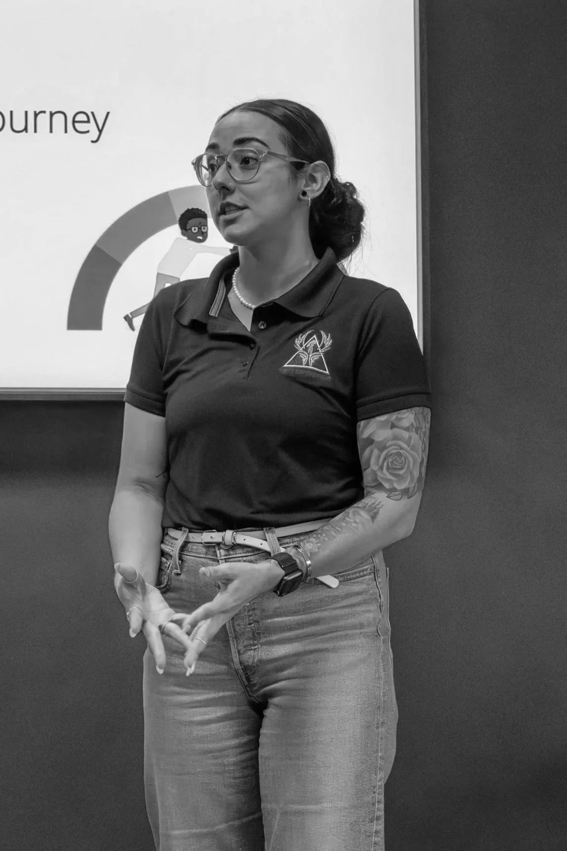 A woman standing in front of a presentation screen, wearing glasses, a polo shirt with a logo, jeans, and a watch, with a tattooed arm and pearl necklace.