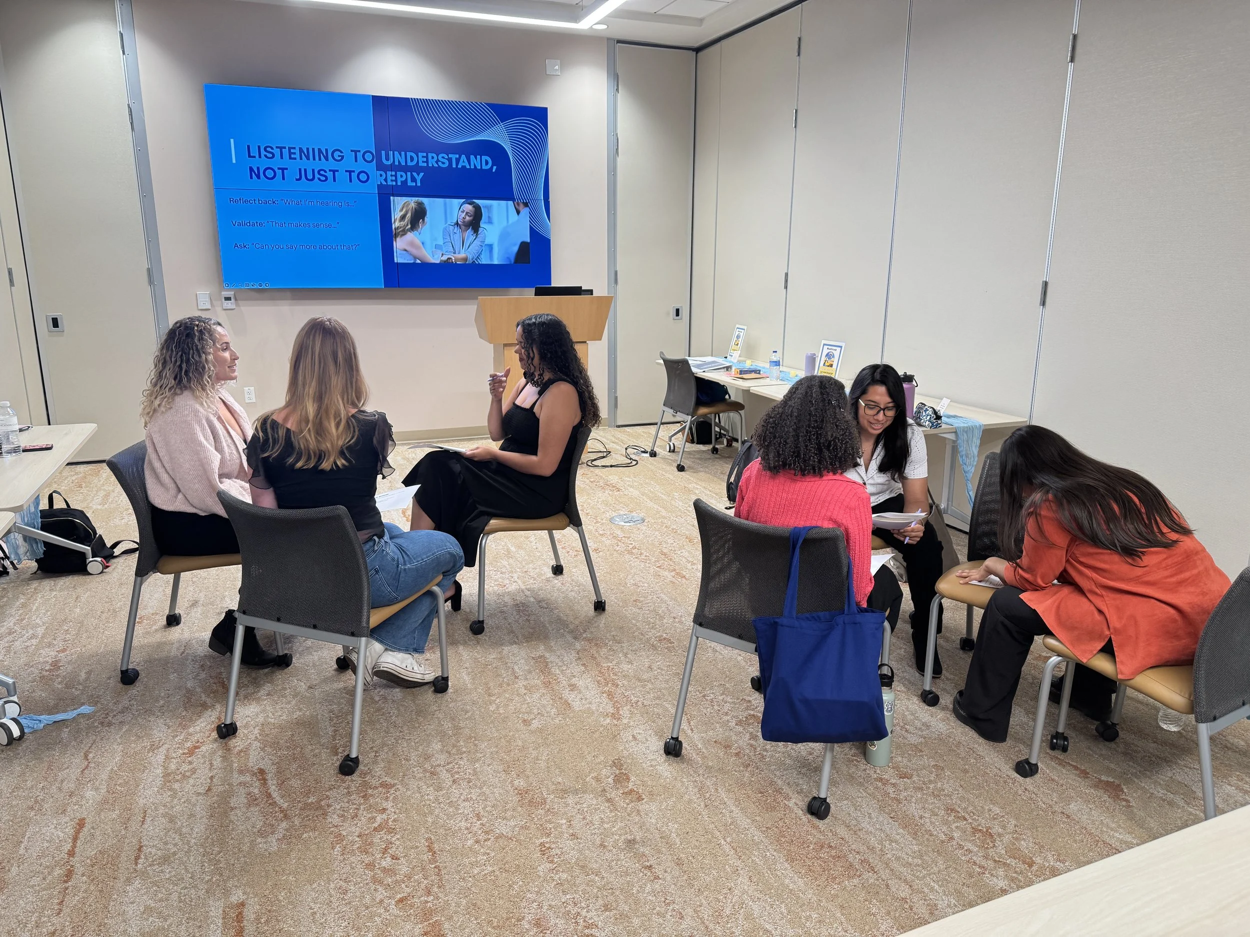 A group of six women sitting in small circles on chairs and engaging in conversation in a conference room with a large digital screen on the wall displaying a presentation about listening and understanding.