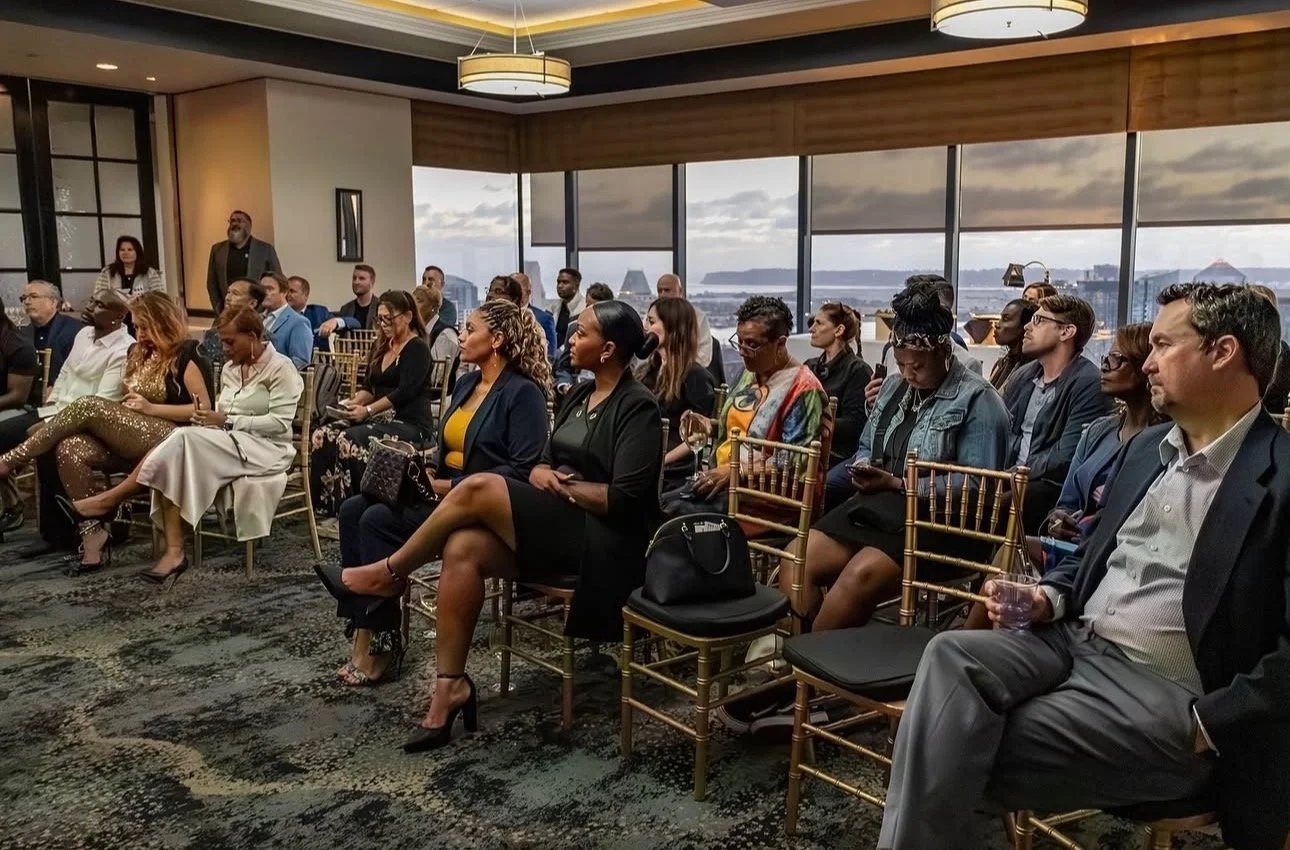 People attending a formal event in a high-rise conference room with large windows, city and water views, sitting on gold chairs, some using phones, some listening attentively.